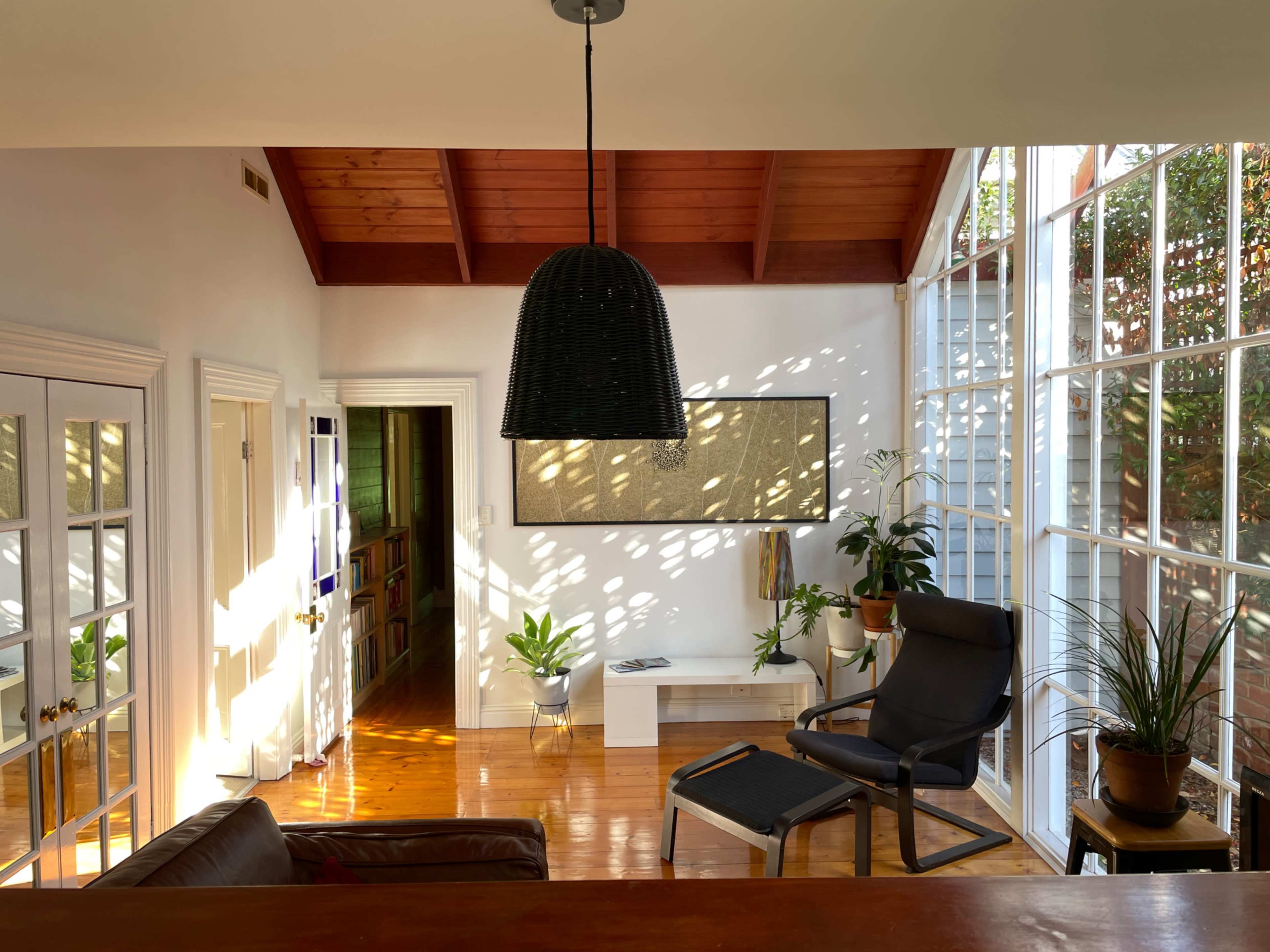 A sunlit room features a black pendant light, a lounge chair, potted plants, and large windows, with wooden ceiling beams overhead.