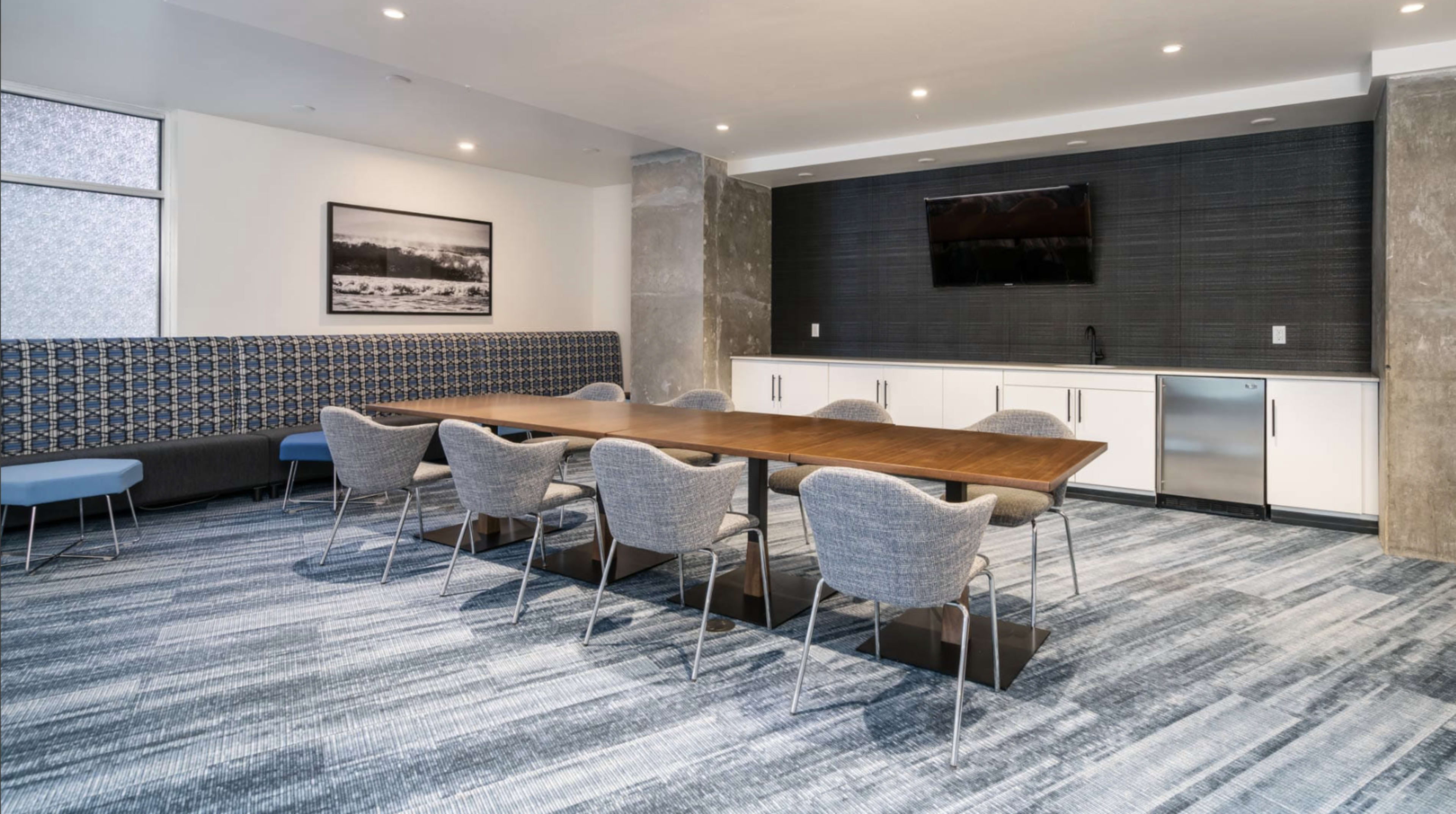 The image shows a modern conference room with a long wooden table surrounded by gray chairs, a kitchenette area, and large windows providing natural light.