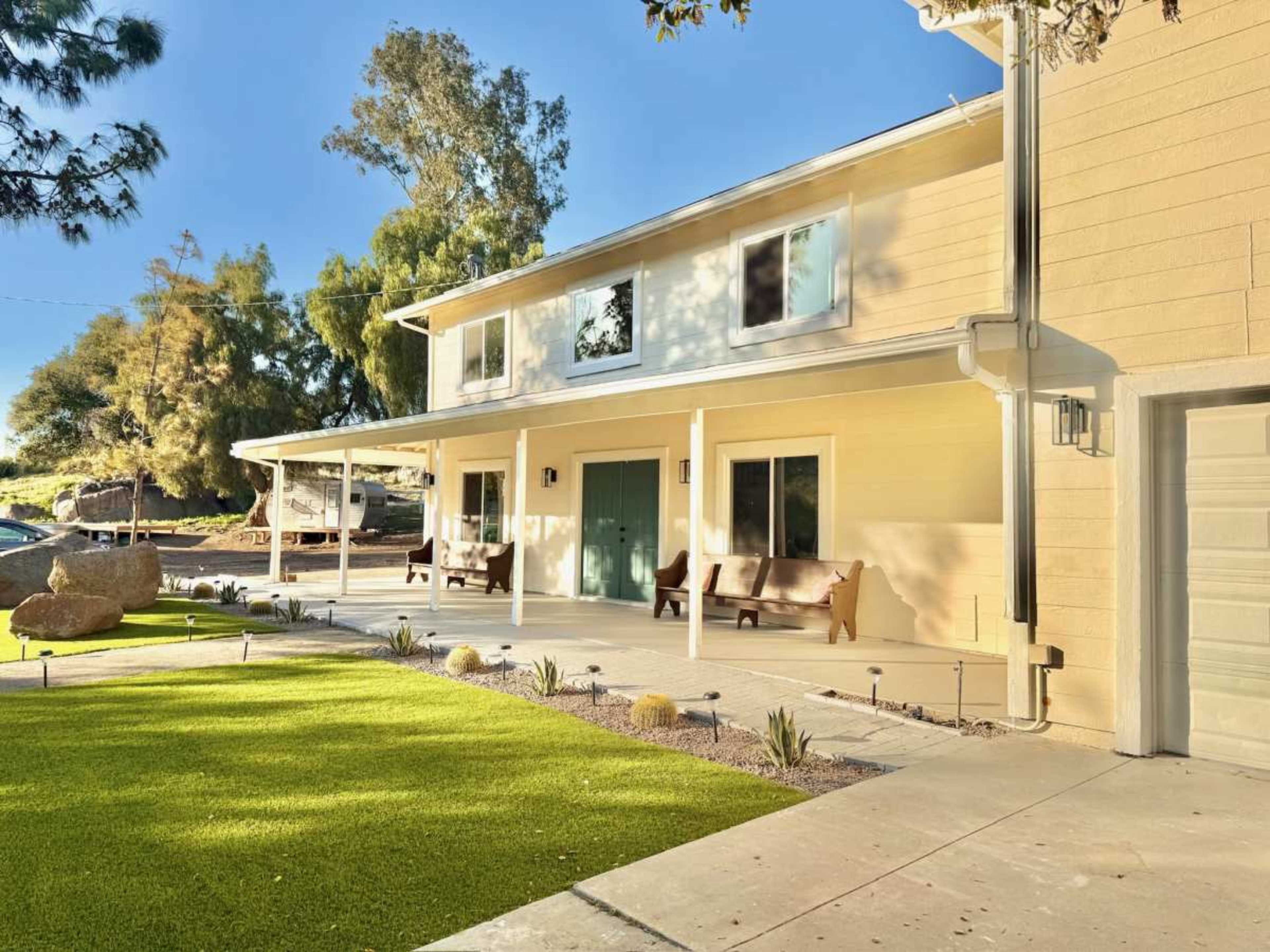 The image shows a two-story house with a porch, surrounded by green grass and various plants.
