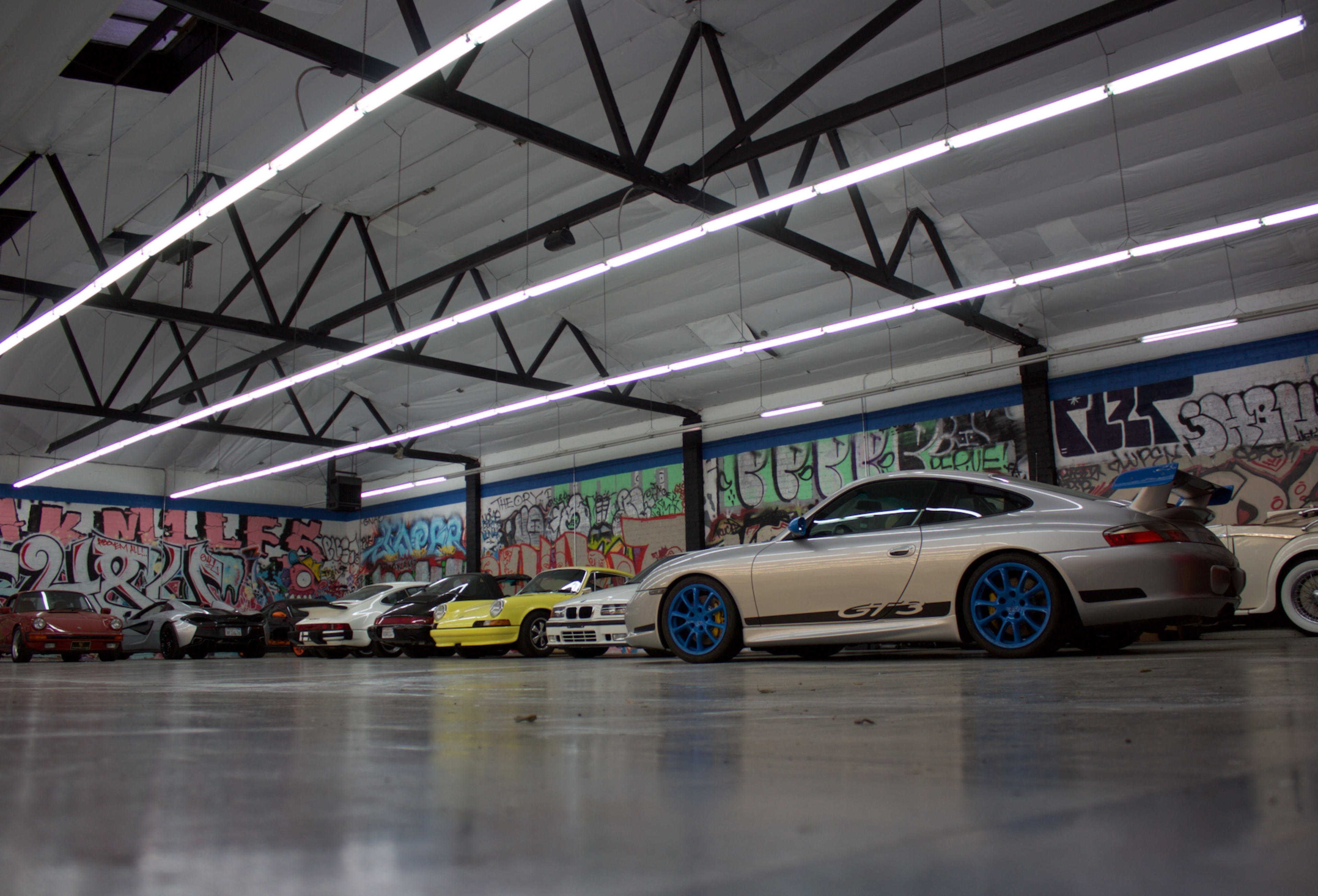 A row of various cars, including a Porsche with blue wheels, is parked in an industrial space adorned with colorful graffiti.