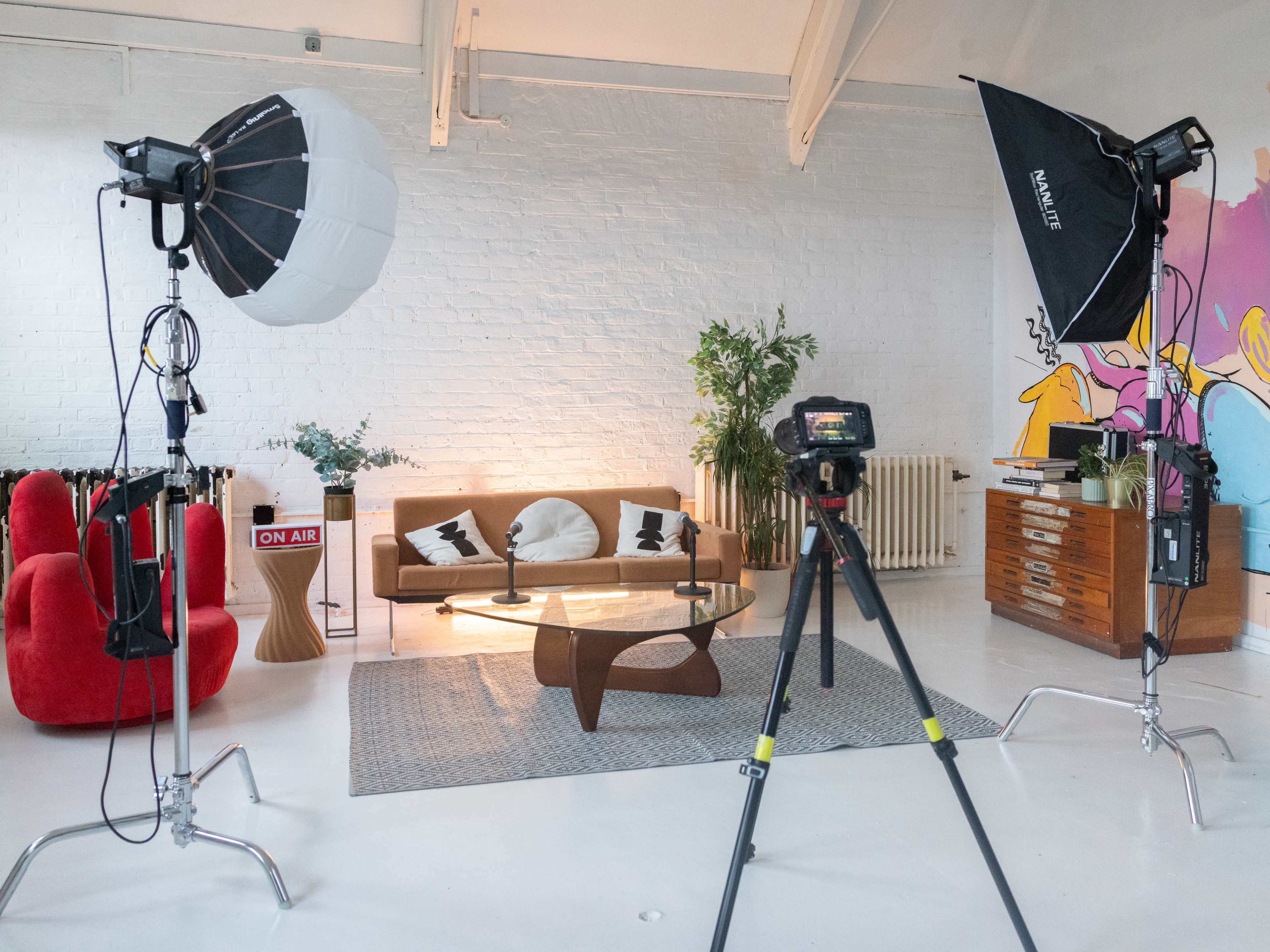 The image shows a well-lit studio space with a sofa and coffee table in the center, flanked by photography equipment, including softbox lights and a camera on a tripod.