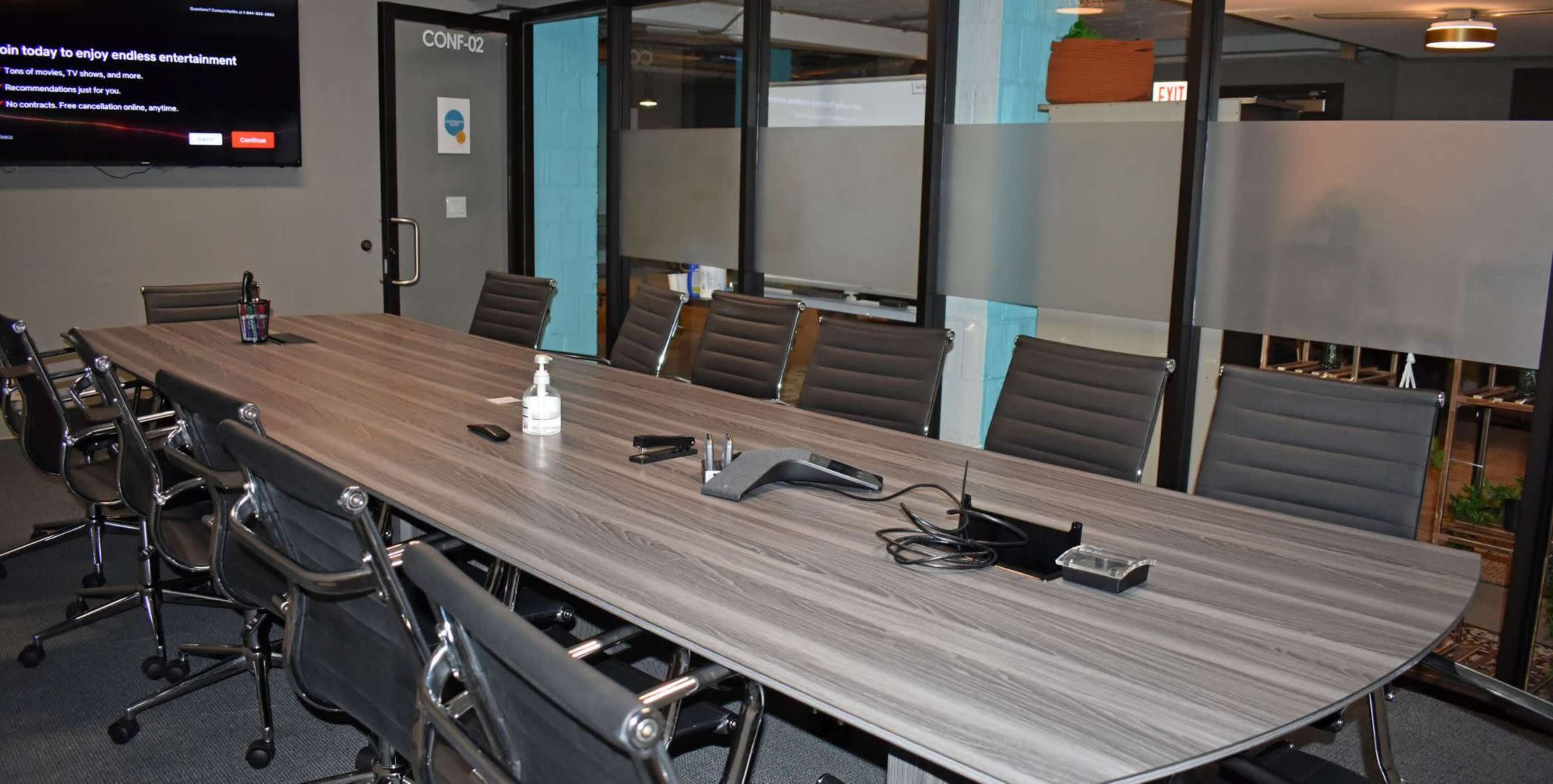 A large conference room features a long wooden table surrounded by black rolling chairs, equipped with a phone and various office supplies, and a screen displaying information on the wall.