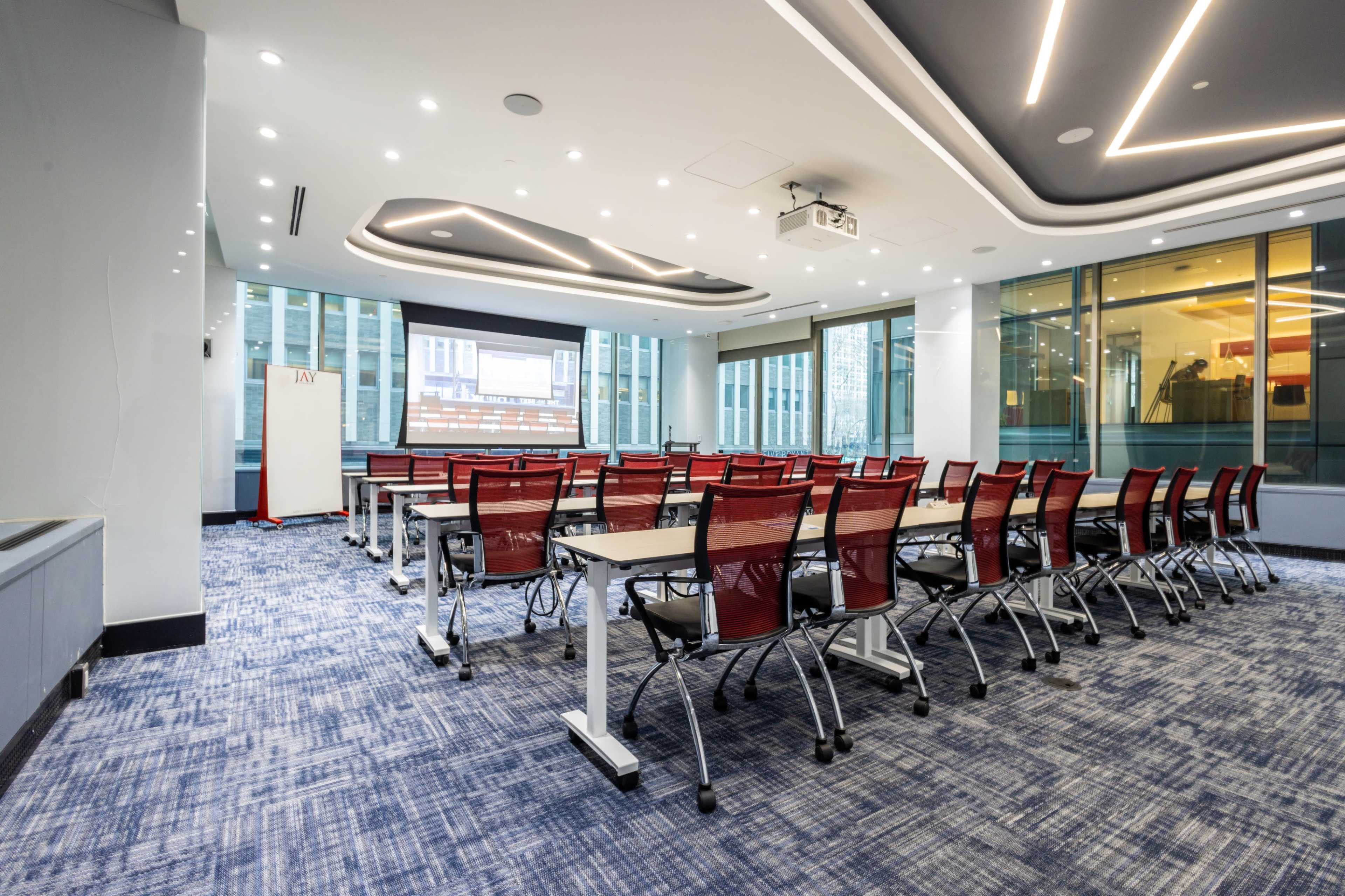 A modern conference room features rows of desks with red chairs facing a projector screen, illuminated by bright overhead lights.