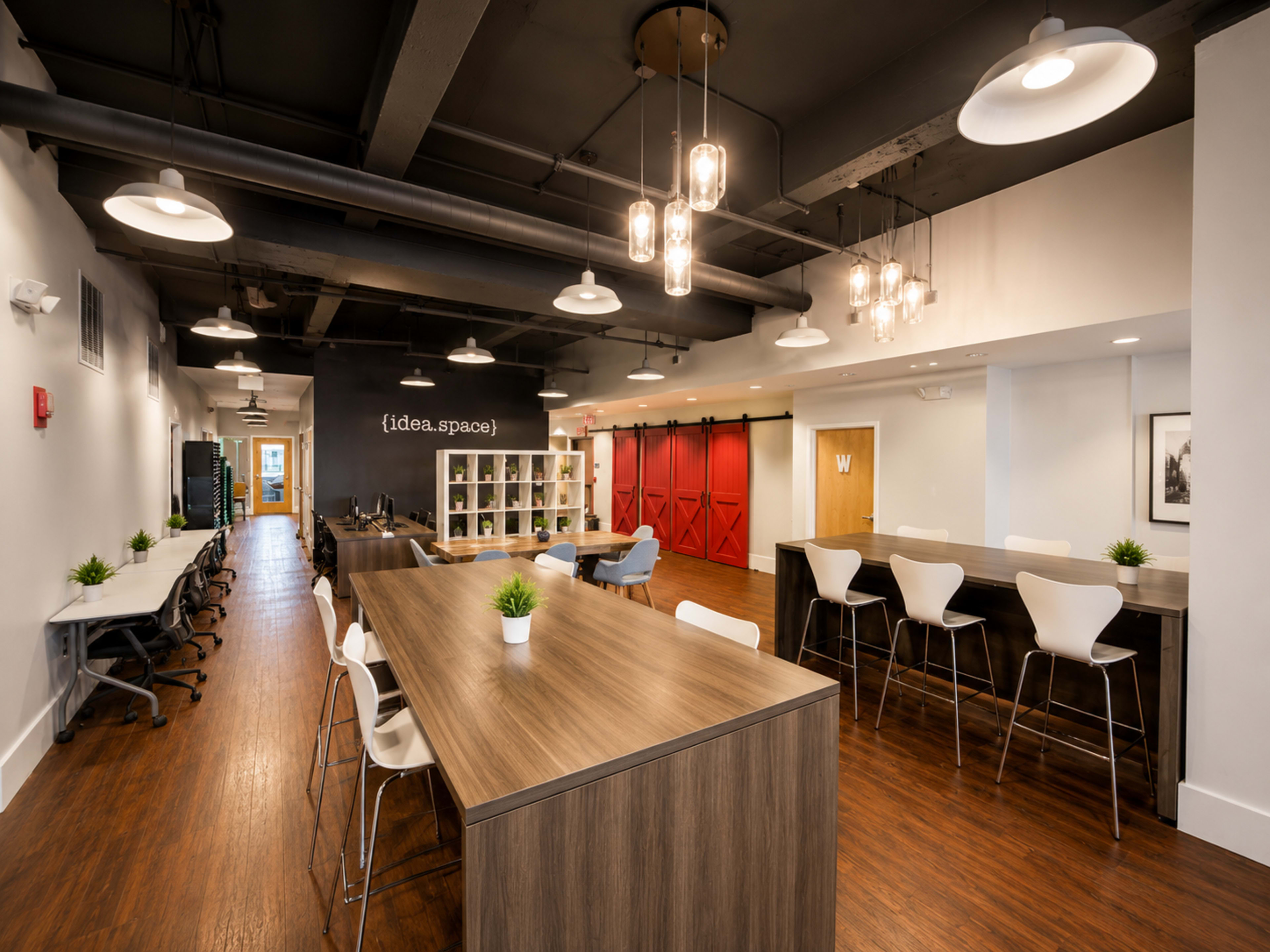 The image shows a modern open office space featuring a long wooden table, contemporary white chairs, and bright overhead lighting, with red sliding doors in the background.