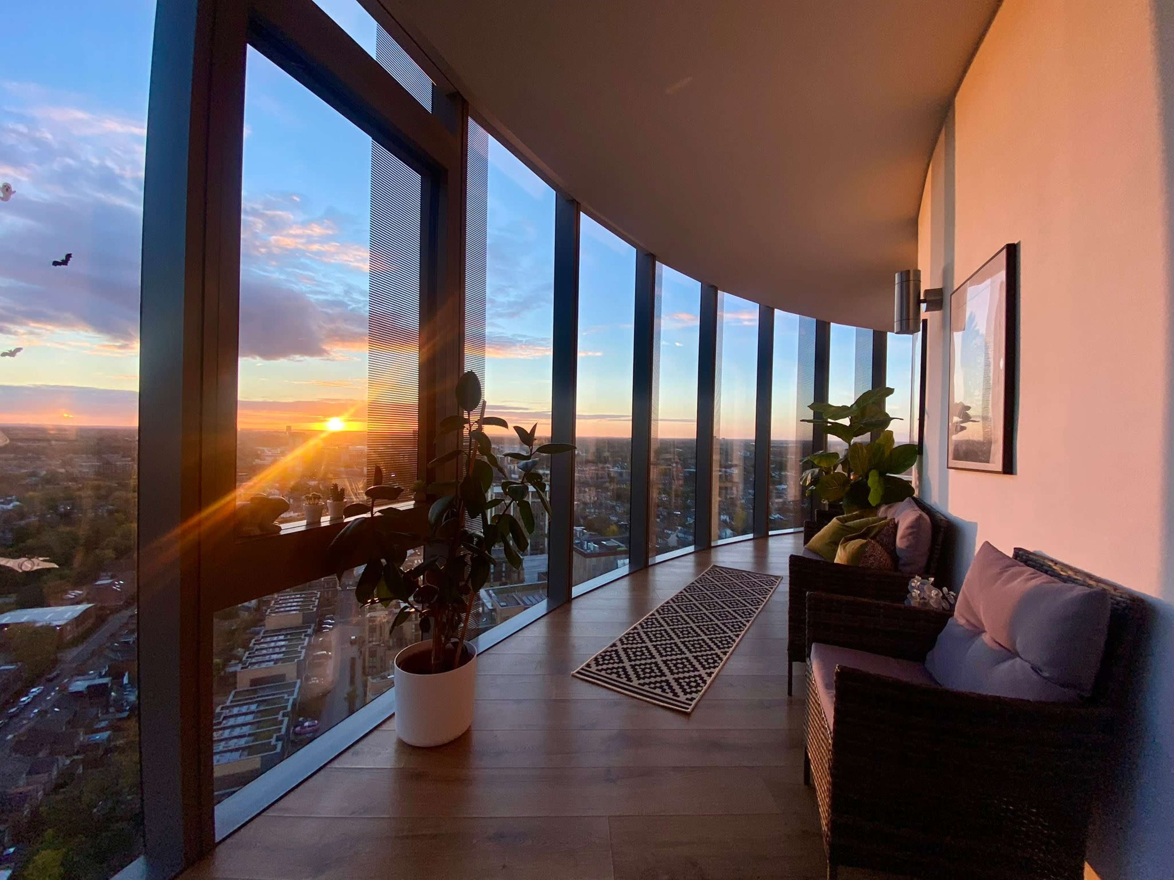 A modern interior with large curved windows, a potted plant, and seating, overlooks a sunset view of a cityscape.