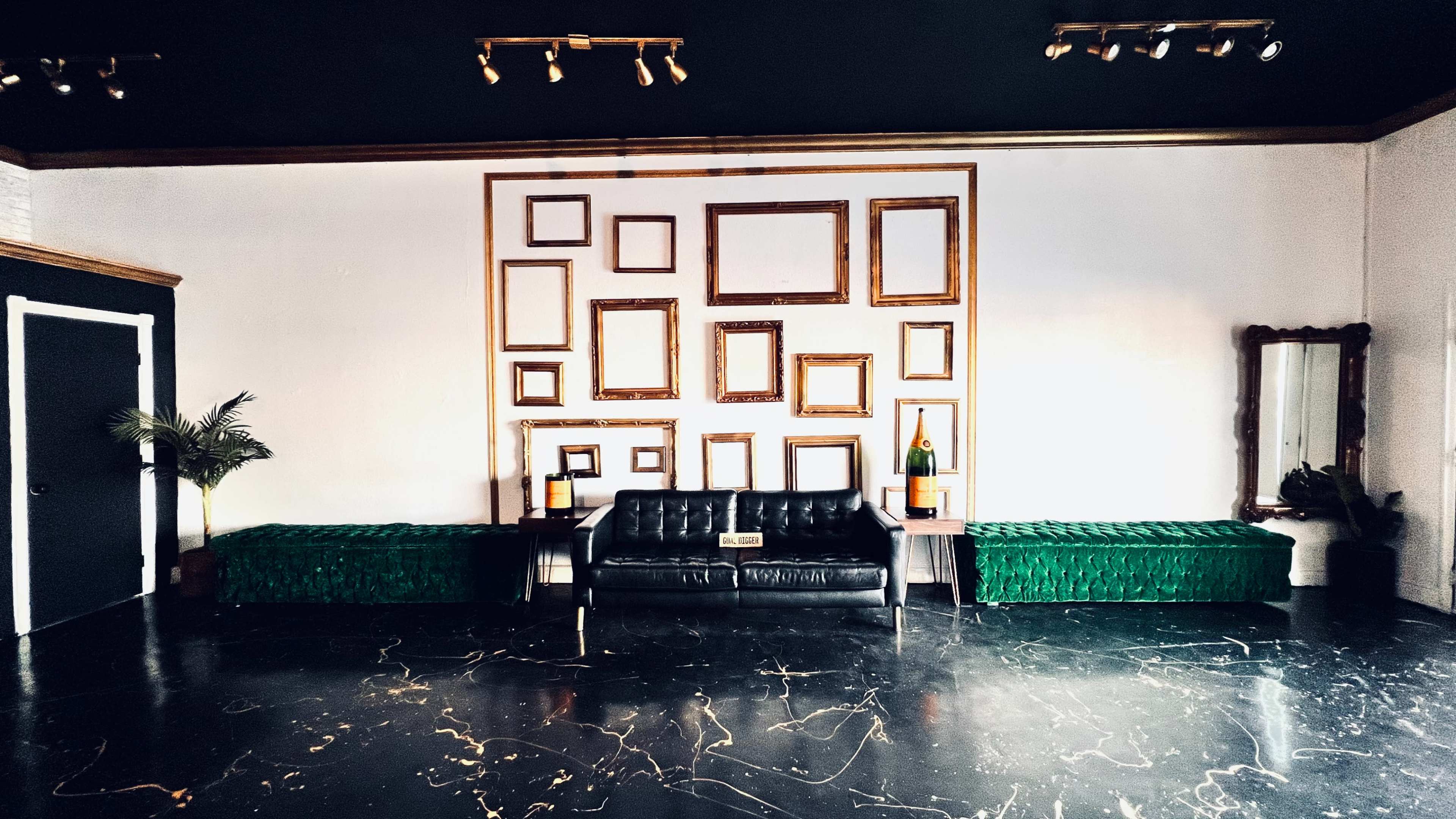 A room with a black leather sofa centered against a wall adorned with various empty gold-framed picture frames, flanked by two green benches.