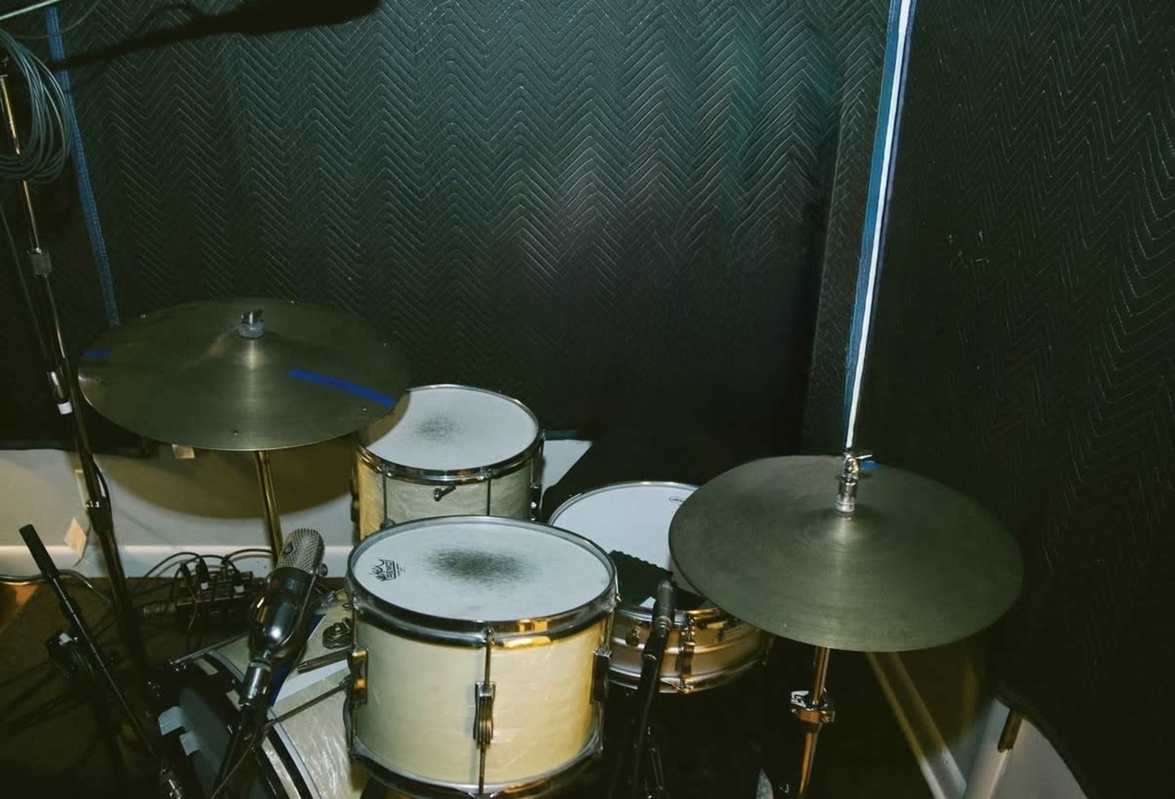 The image shows a drum set positioned in a soundproofed recording space, featuring two cymbals and two drums.