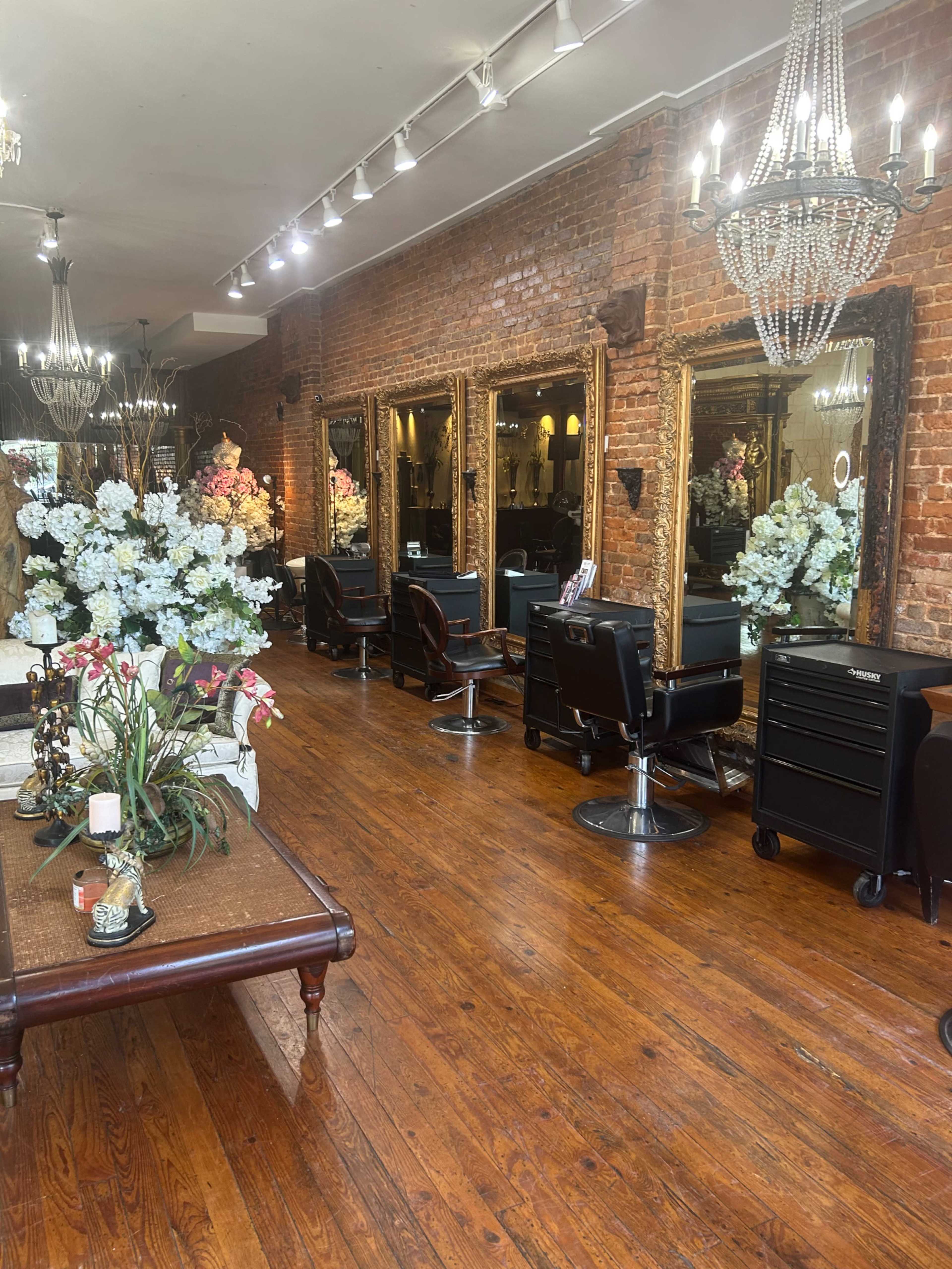The interior of a hair salon features wooden floors, large mirrors in ornate frames, and elegant lighting, with decorated styling stations and floral arrangements.