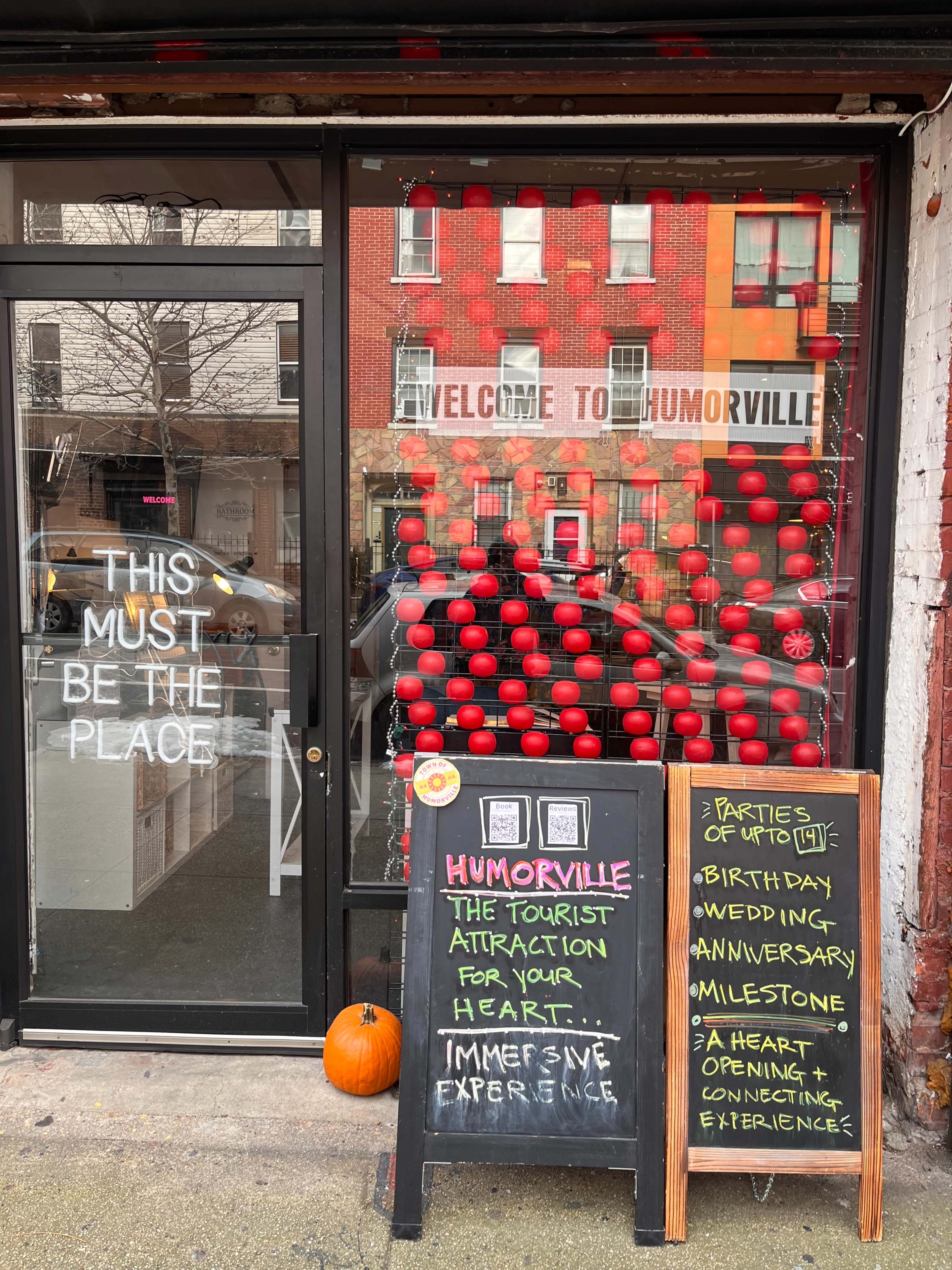 A storefront window displays a vibrant sign reading "WELCOME TO HUMORVILLE" along with red balloons and a chalkboard advertising events and experiences for special occasions.