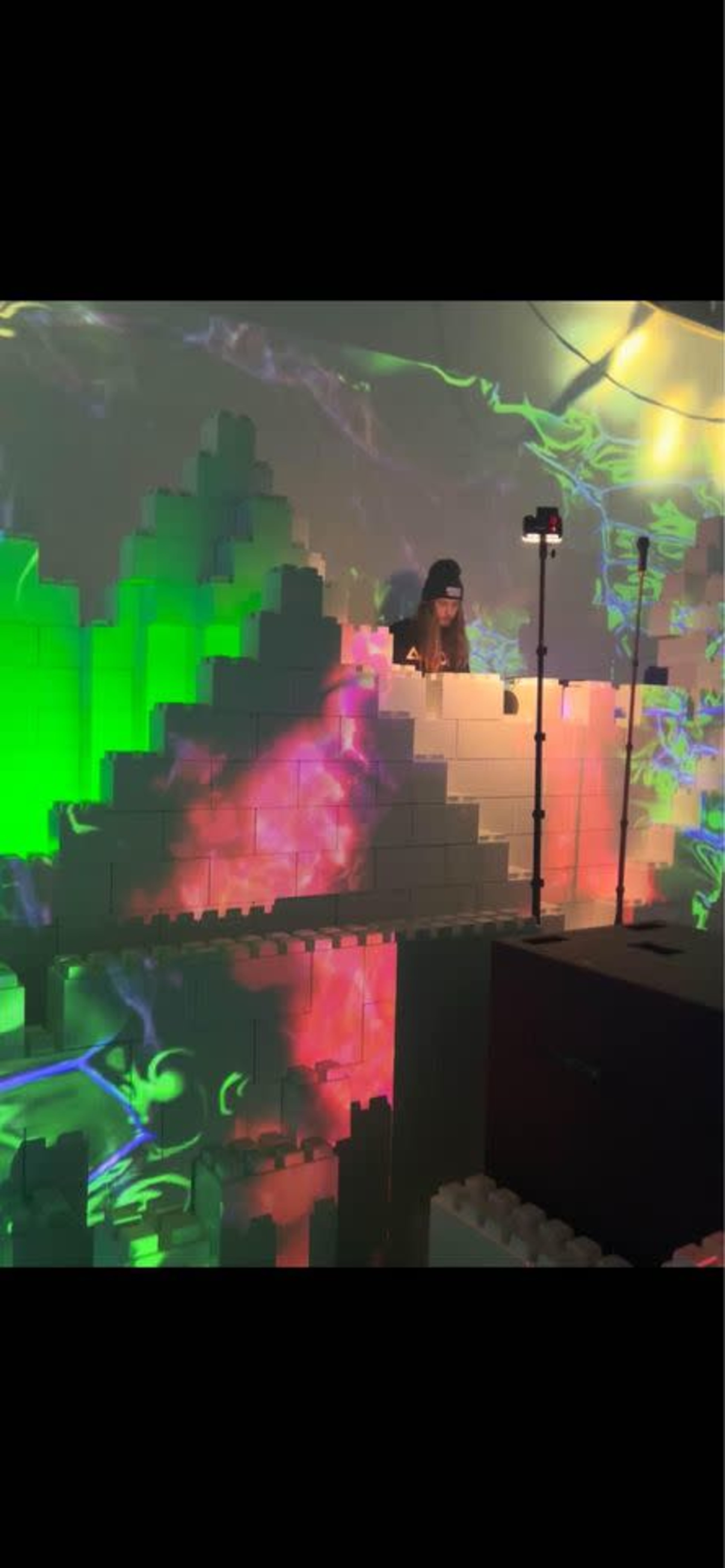 A person operates a DJ setup surrounded by colorful, glowing projections and three-dimensional block structures.