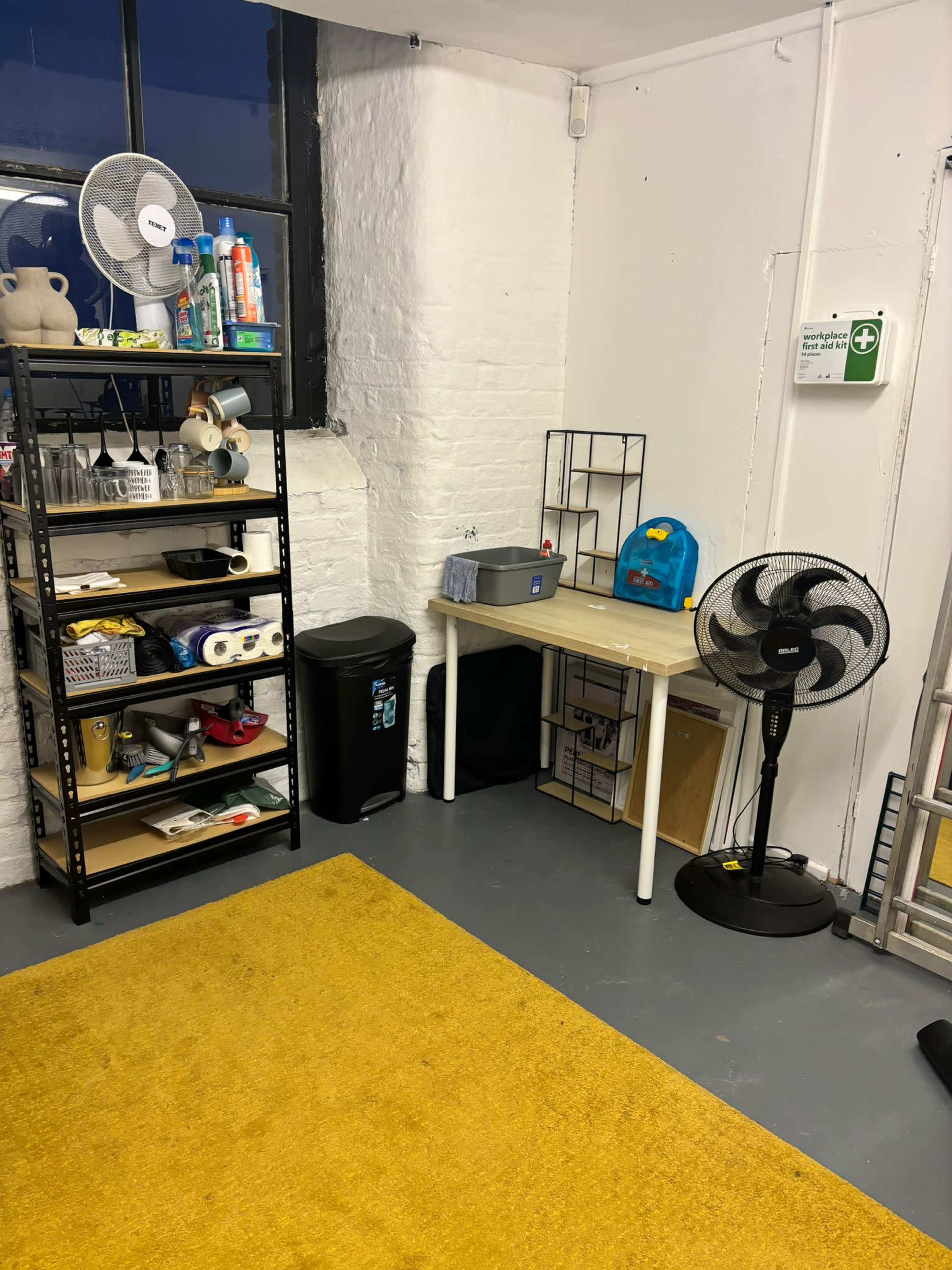 The image shows a tidy room with a yellow rug, shelves containing various items, a small table, a fan, a trash bin, and a blue backpack.