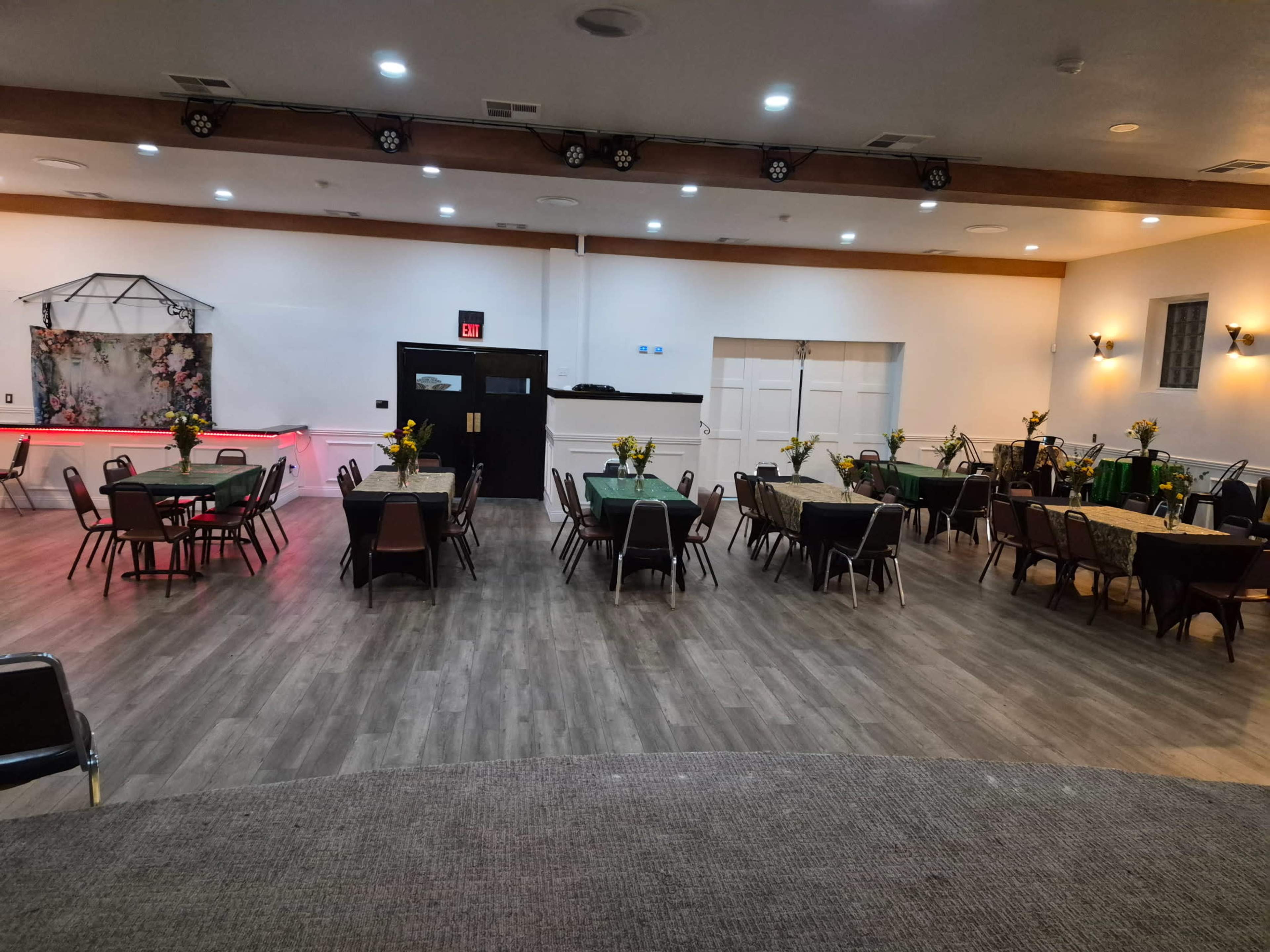 The image shows a spacious event hall arranged with multiple tables and chairs, each decorated with yellow flowers, and featuring an illuminated backdrop.