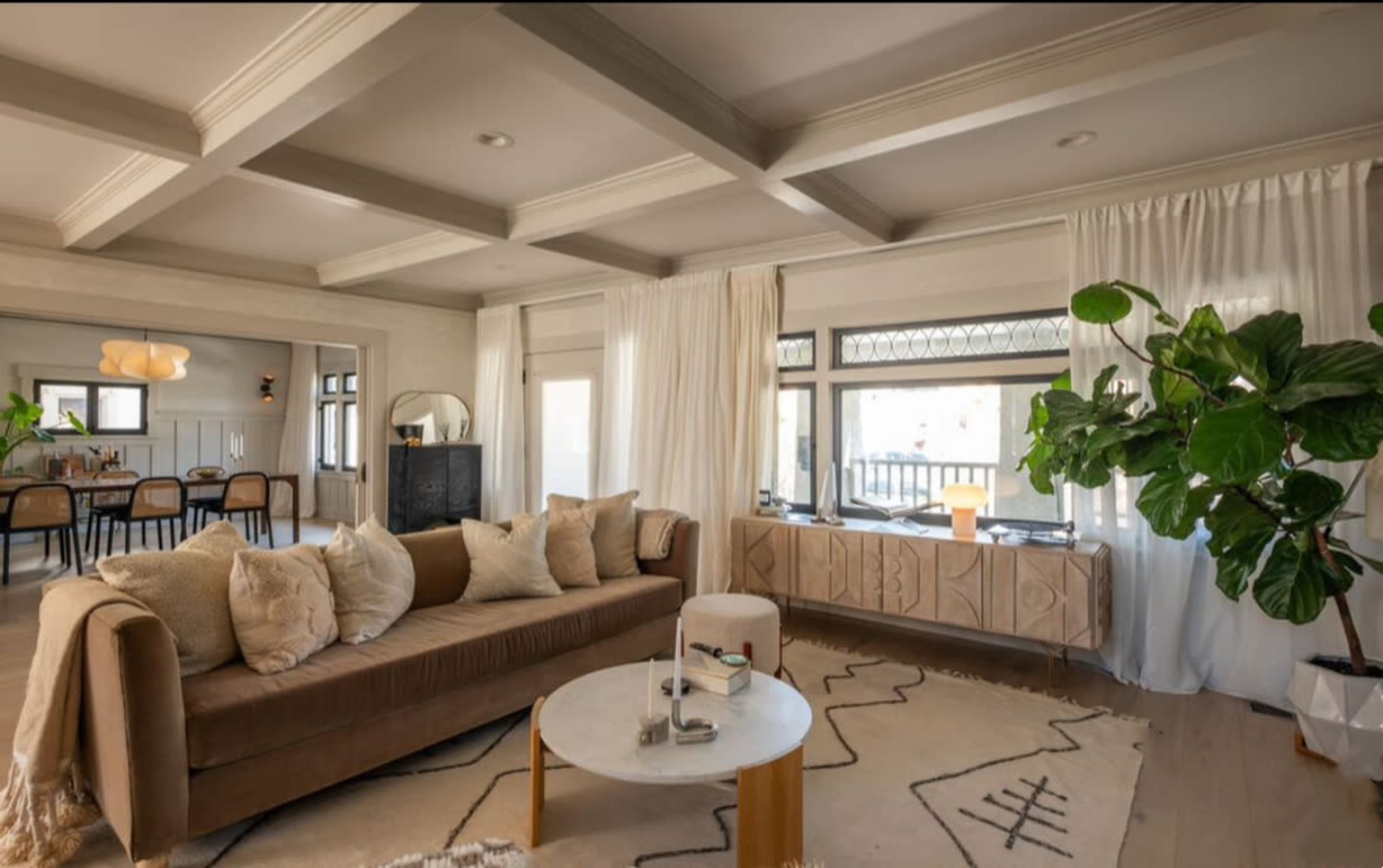 The image shows a modern living room featuring a brown sofa, a round coffee table, large windows with drapes, and a potted plant, with a dining area visible in the background.