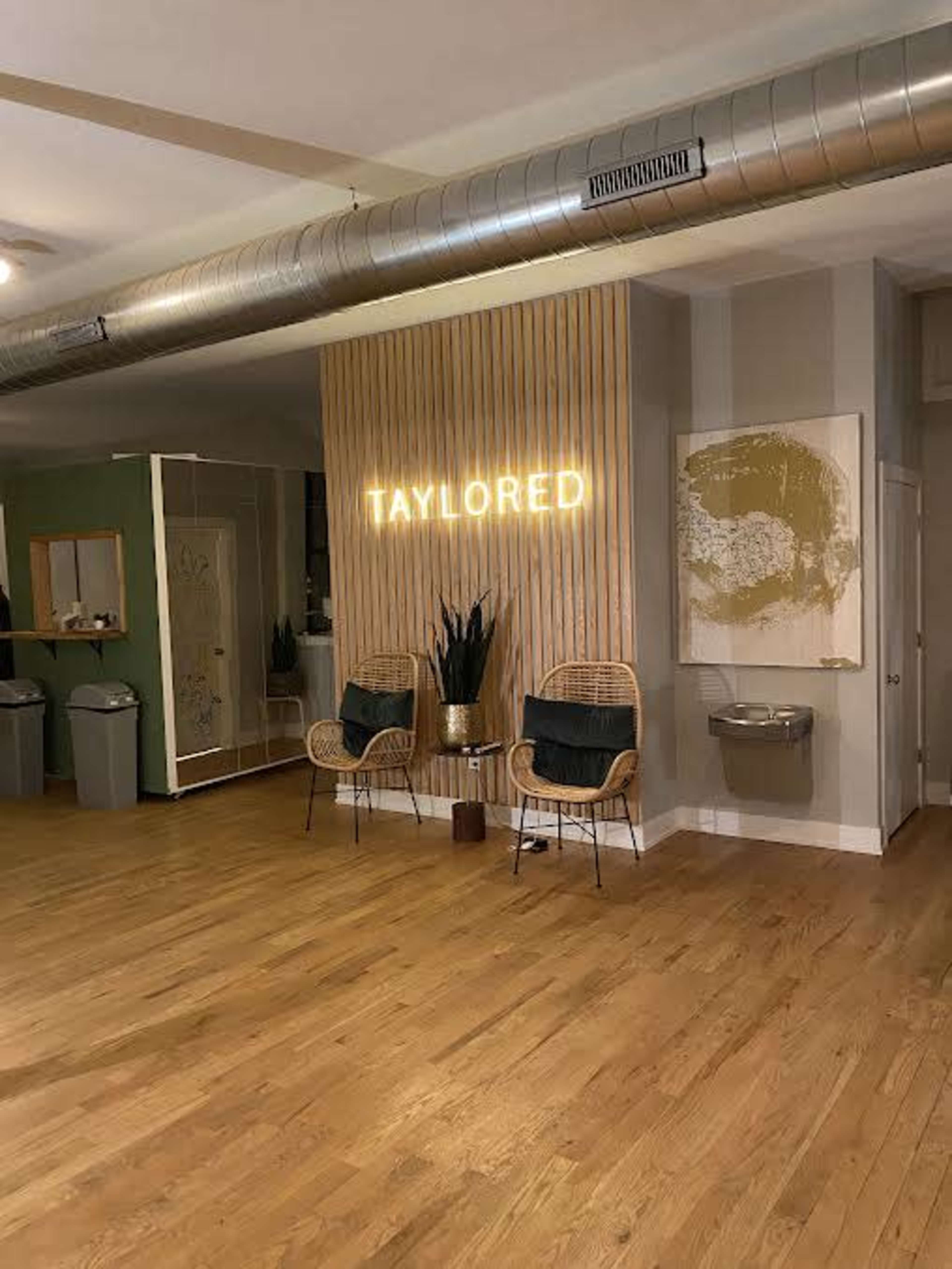 A spacious room with wooden floors, featuring a neon sign that reads "TAYLORED," two rattan chairs, and a small table, along with a water fountain and decorative plants.