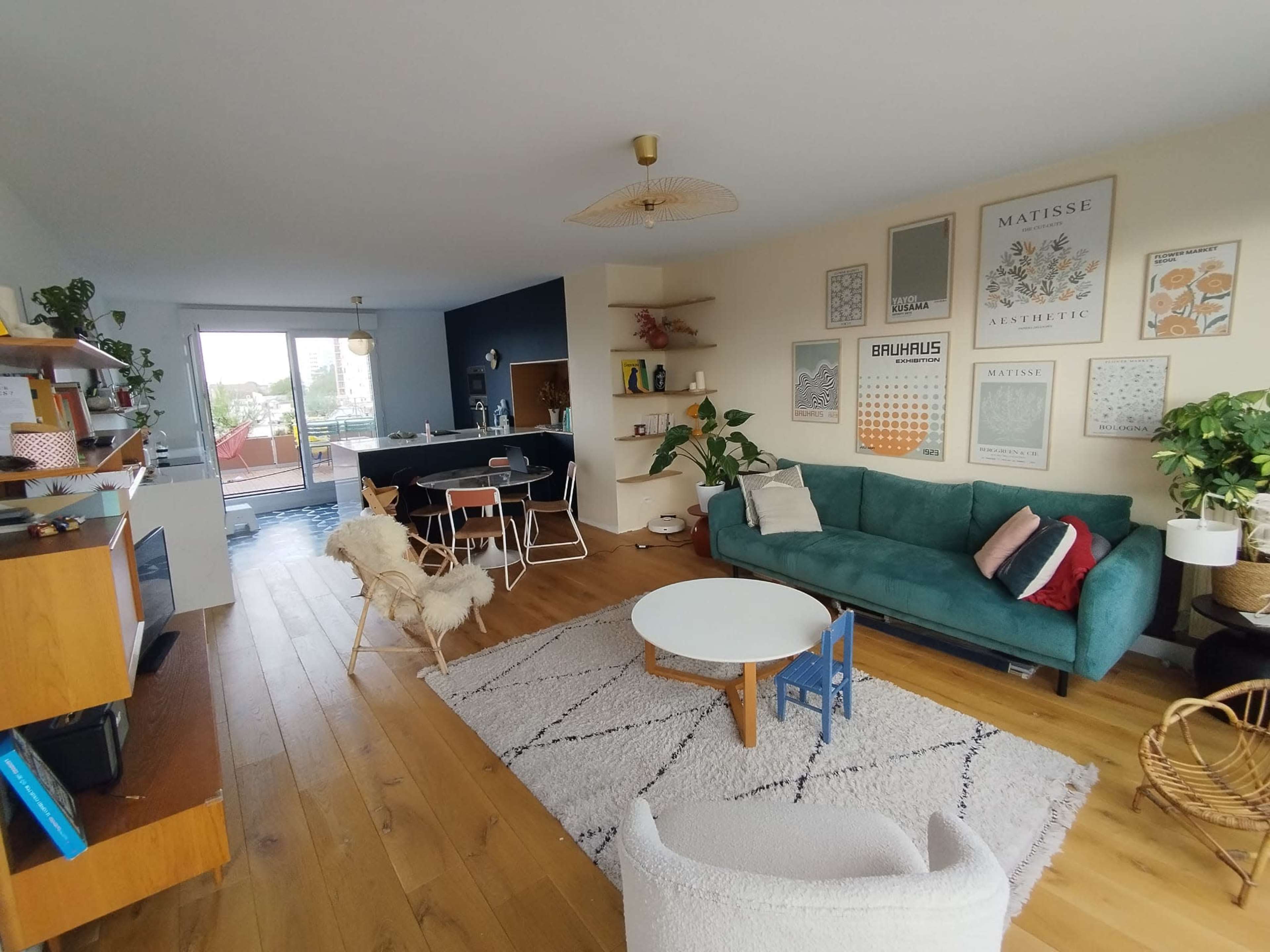 The living room features a green sofa, a round coffee table, wooden flooring, and wall decorations, with a view of an outdoor space visible through a large window.