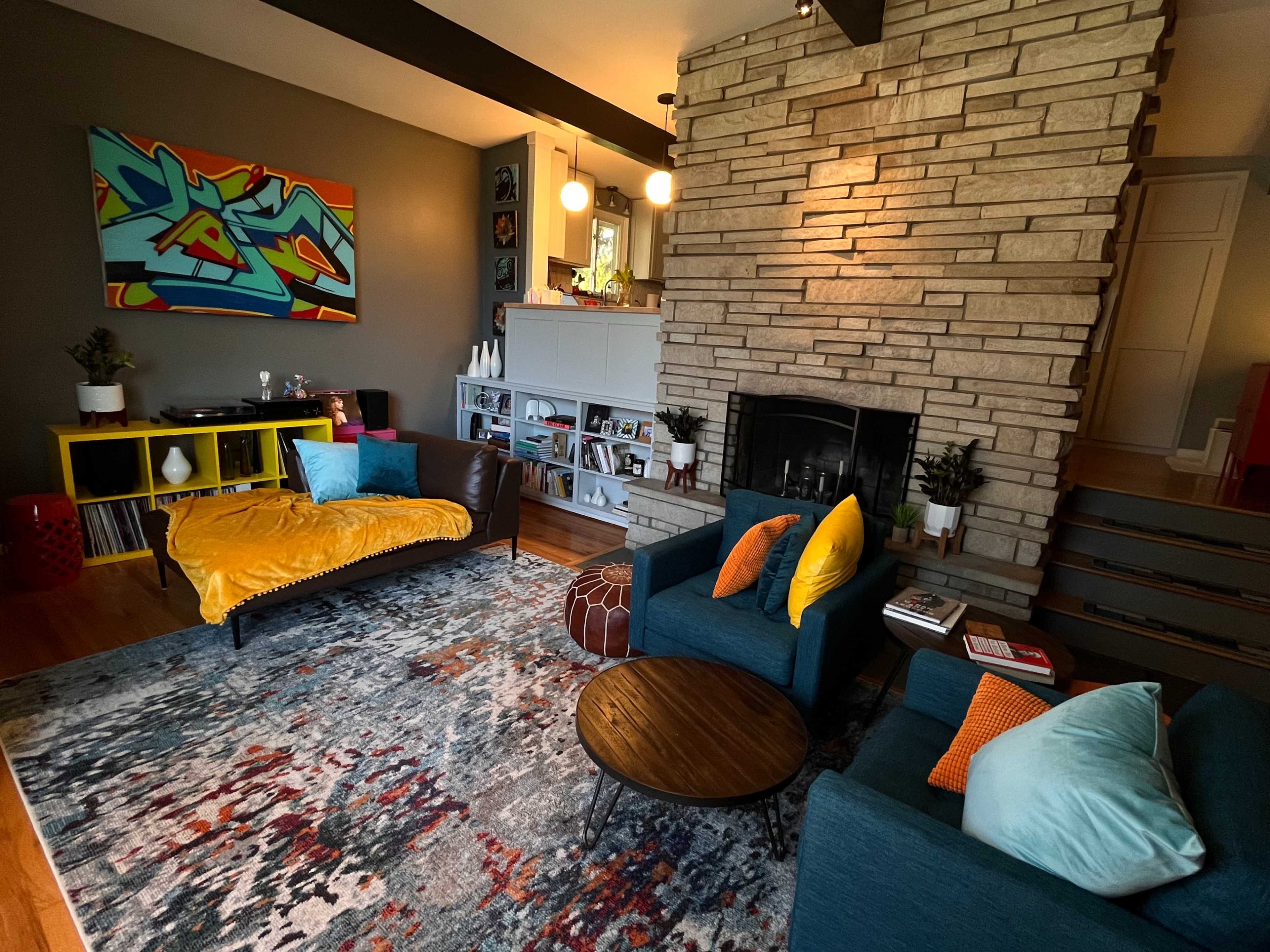 A modern living room featuring a stone fireplace, a colorful area rug, a yellow couch, and blue armchairs with decorative pillows.