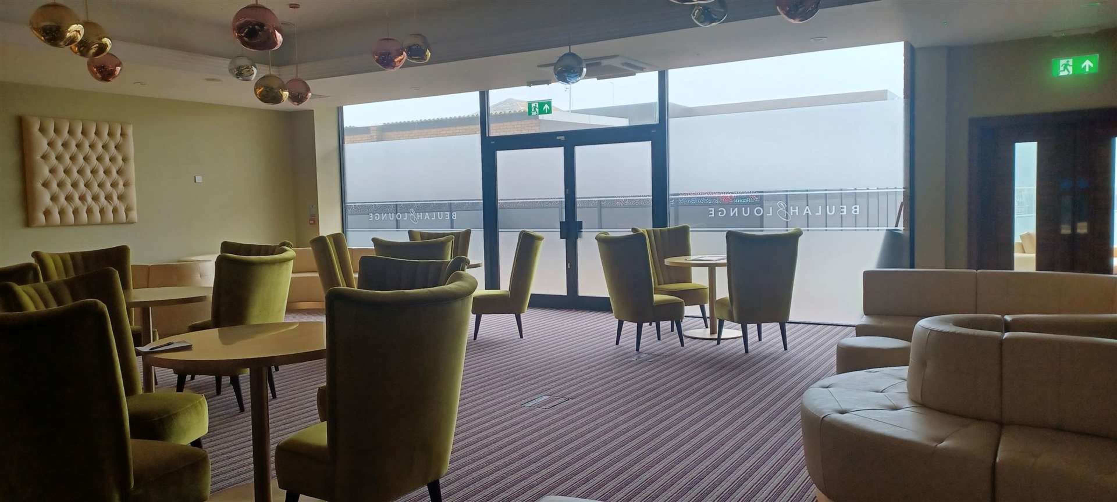 A modern lounge area with green upholstered chairs, circular tables, and a large glass entrance with frosted panels.