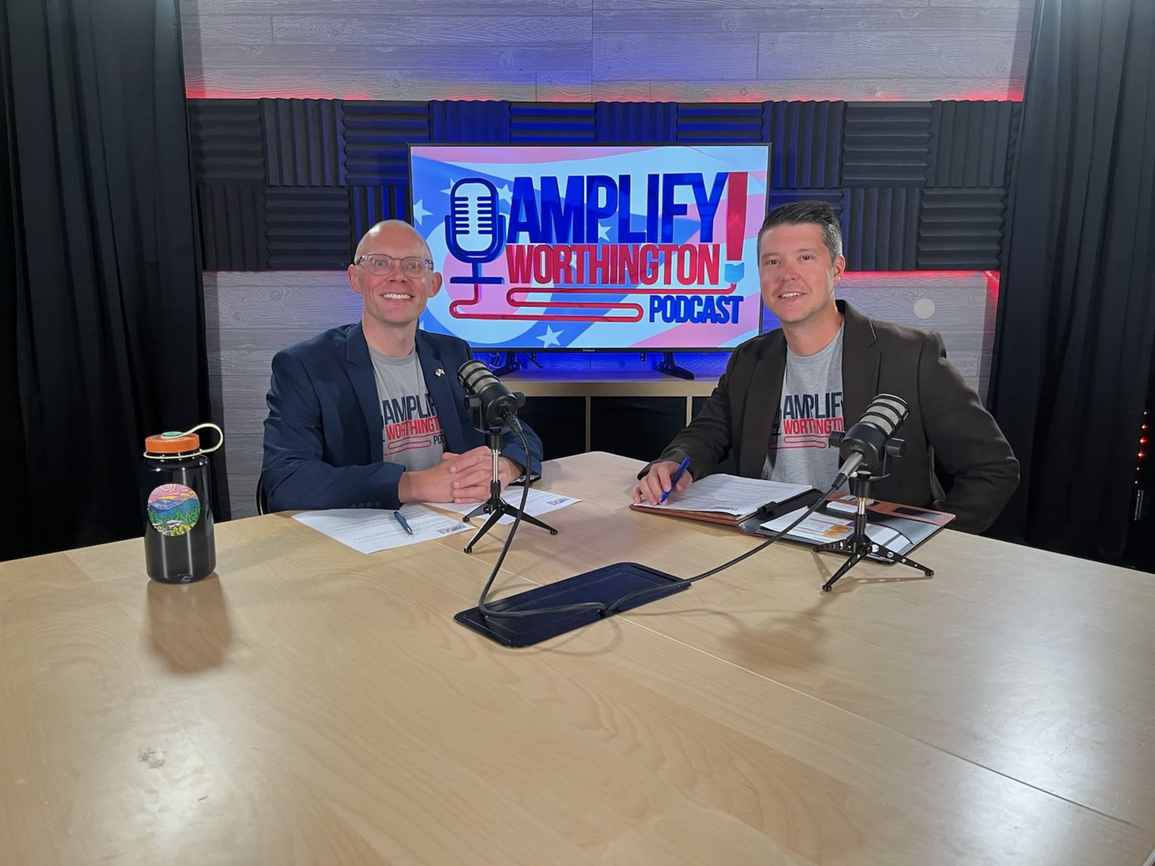 Two men sit at a table with microphones in front of them, preparing for a recording of the "Amplify Worthington" podcast, with a backdrop displaying the podcast's logo.