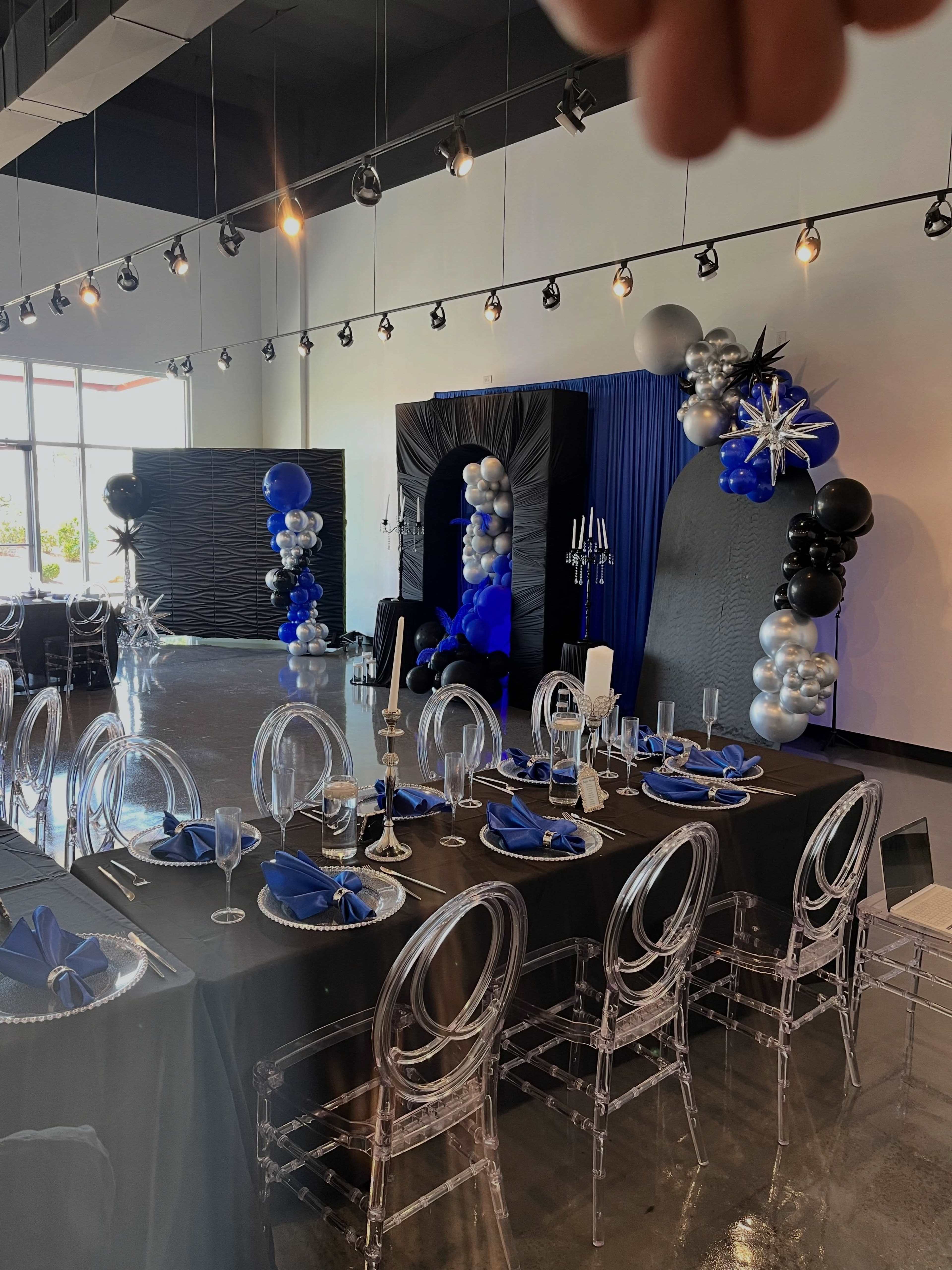 The image shows a dining setup in a modern venue with a black and blue color scheme, featuring clear acrylic chairs, elegant table settings, and decorative balloon arrangements.