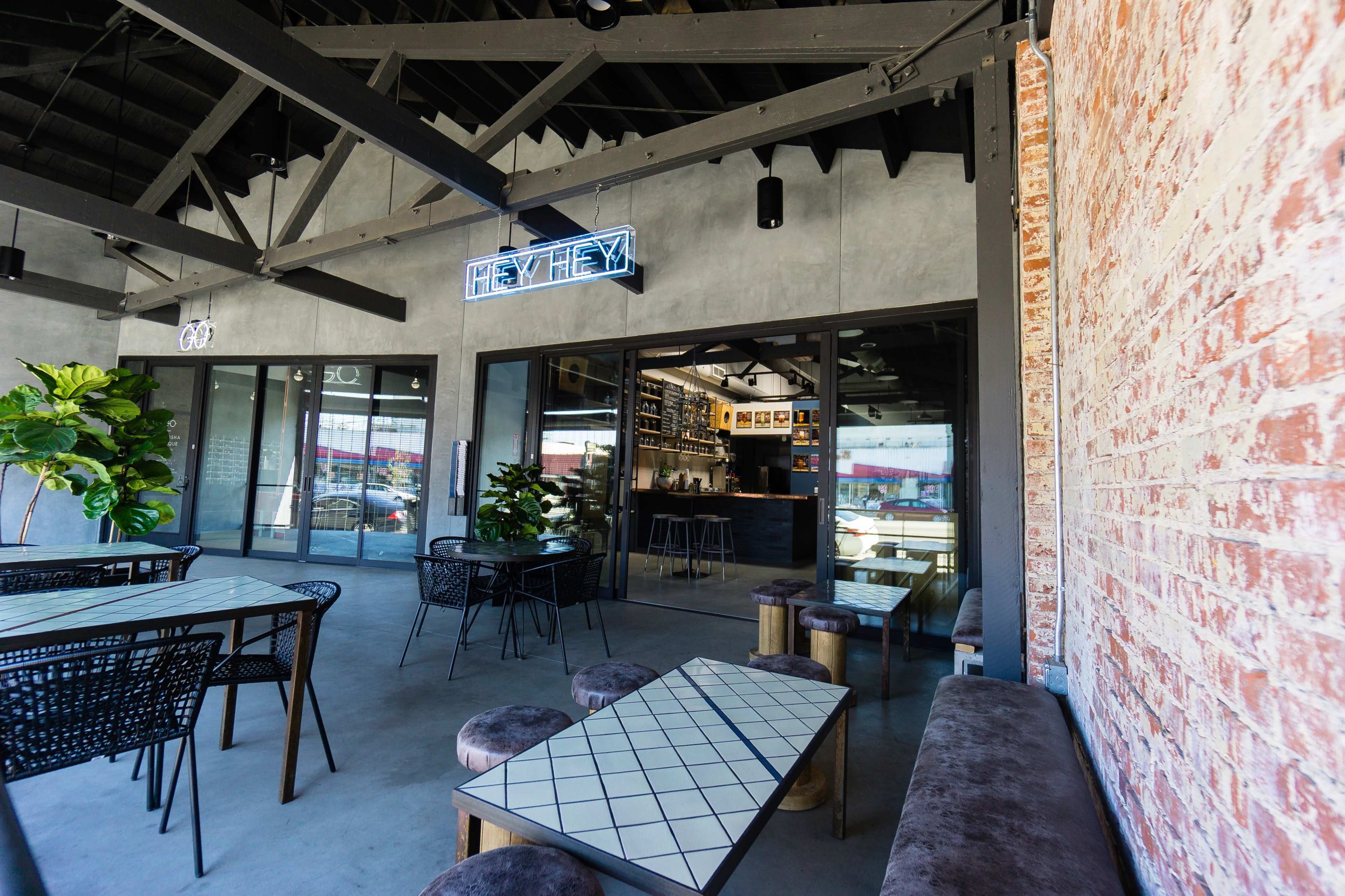 A modern café with a brick wall, large windows, and sleek furniture, featuring indoor plants and a bar area in the background.