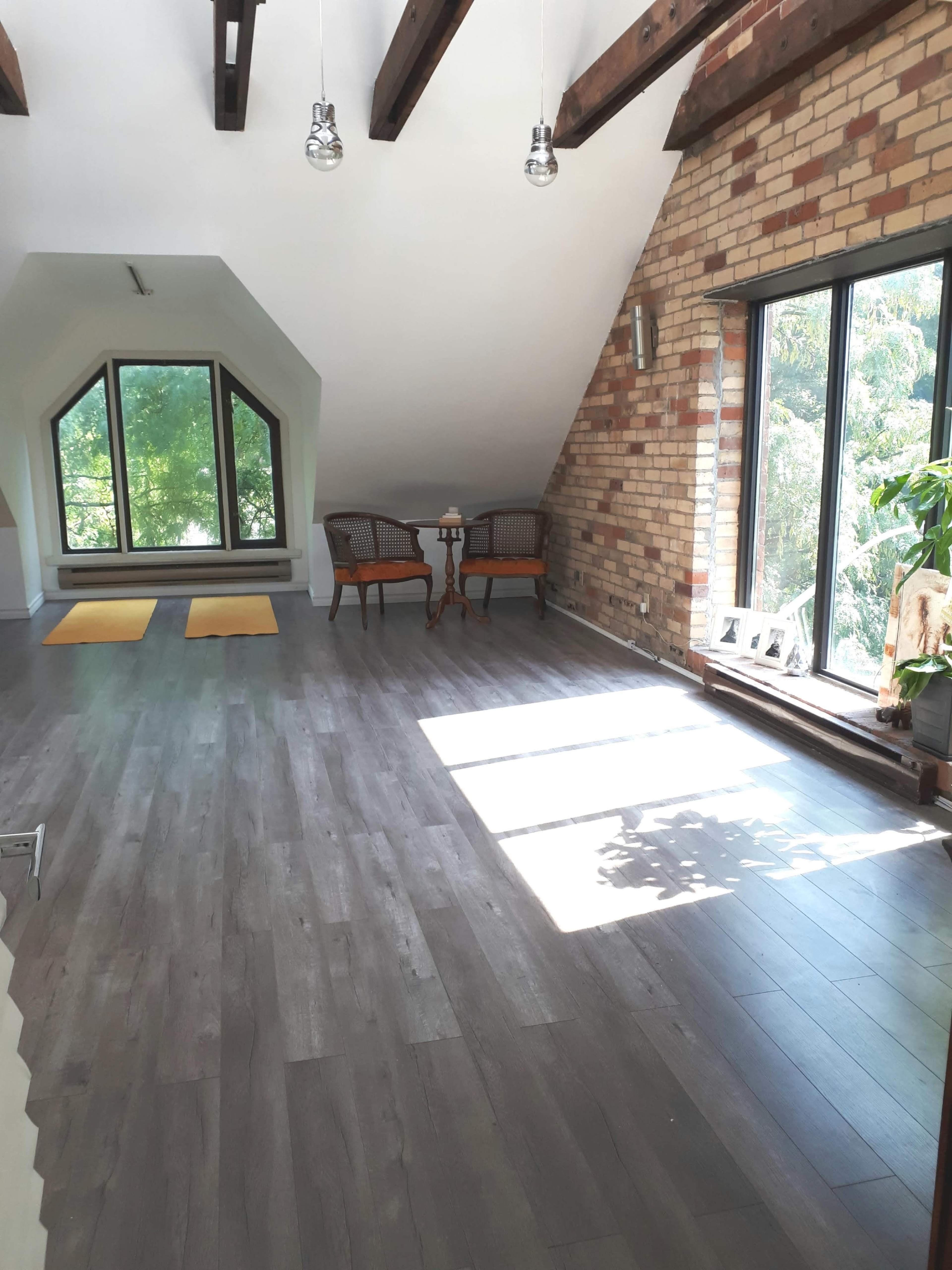 A spacious room with wooden beams, large windows, two chairs, a small table, and yoga mats on the floor.