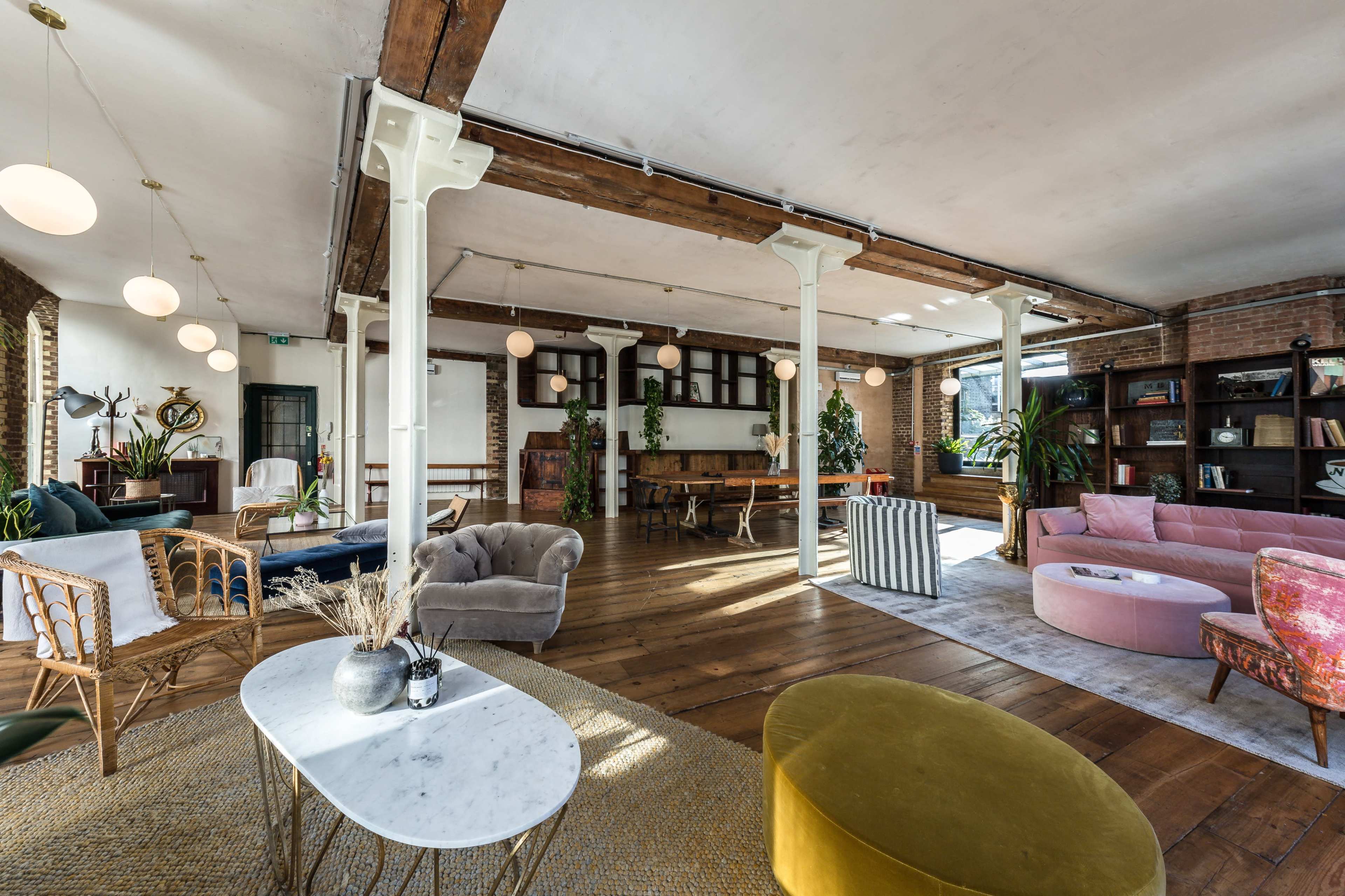 A spacious loft-style interior featuring wooden floors, various seating arrangements, and large windows, adorned with plants and modern light fixtures.
