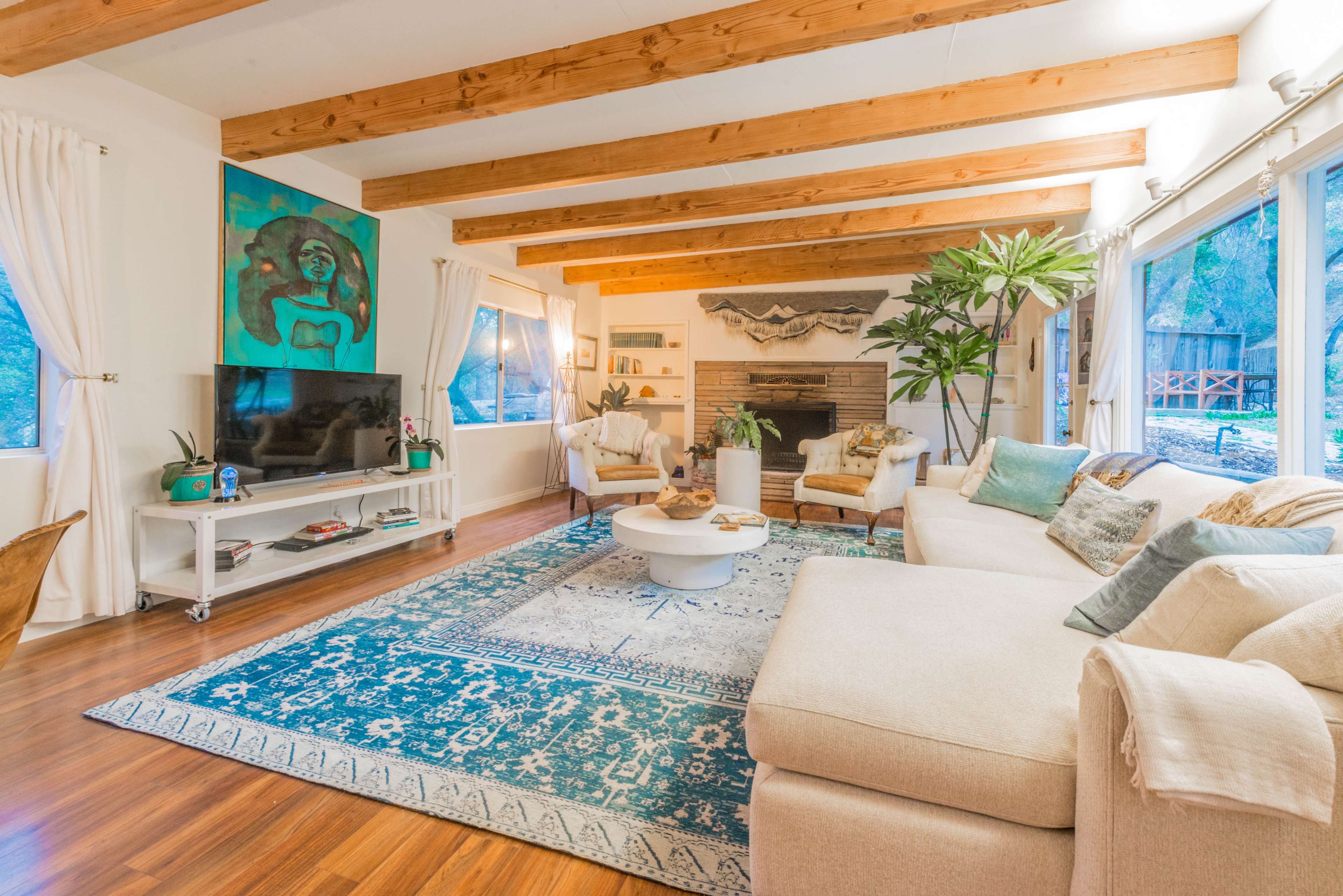 A cozy living room features wooden beams, a sectional sofa, a coffee table, a television, and large windows with views of the outdoors.