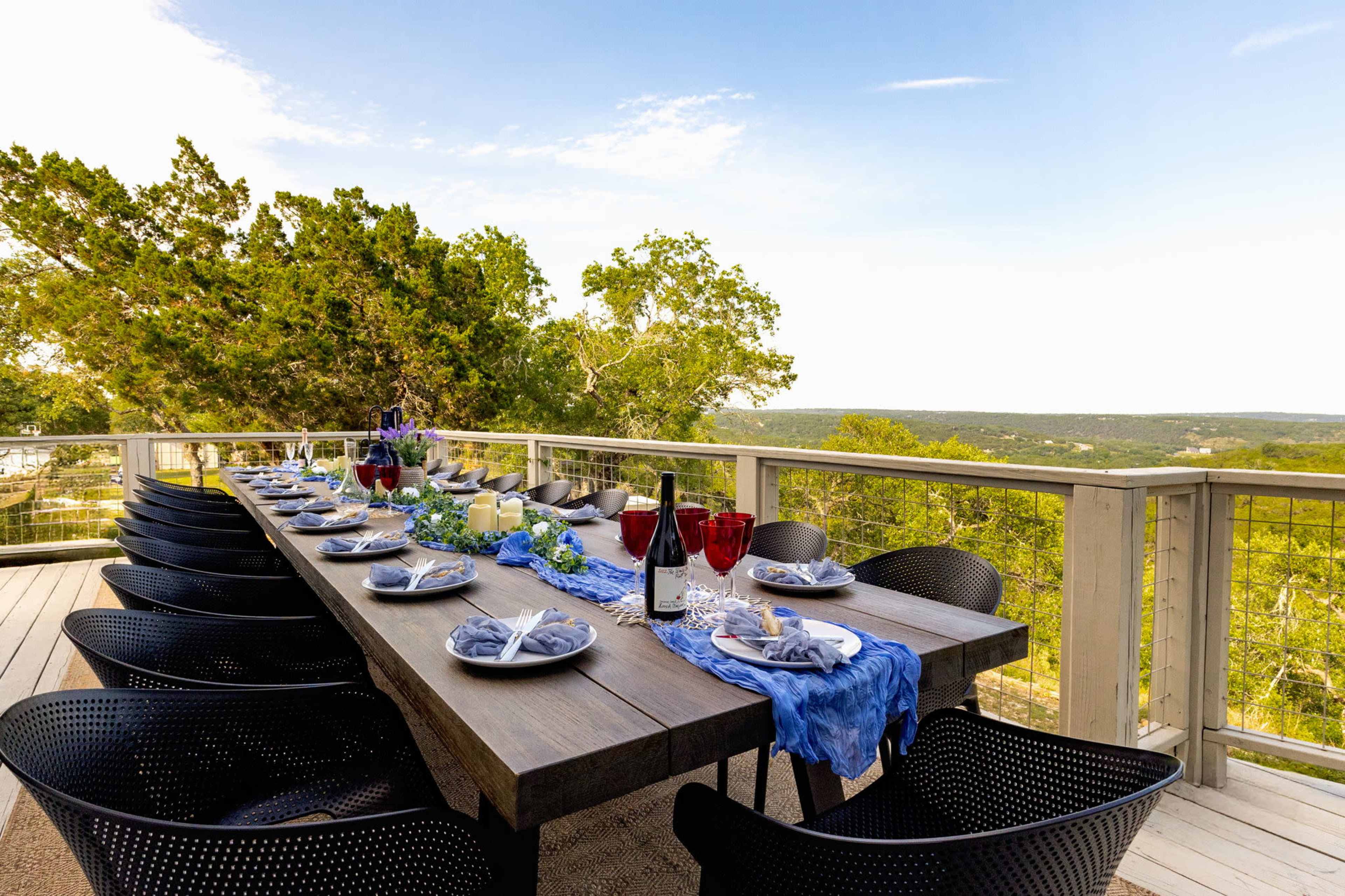 A long dining table is set with plates, glasses, and decorations on a deck overlooking a scenic landscape.