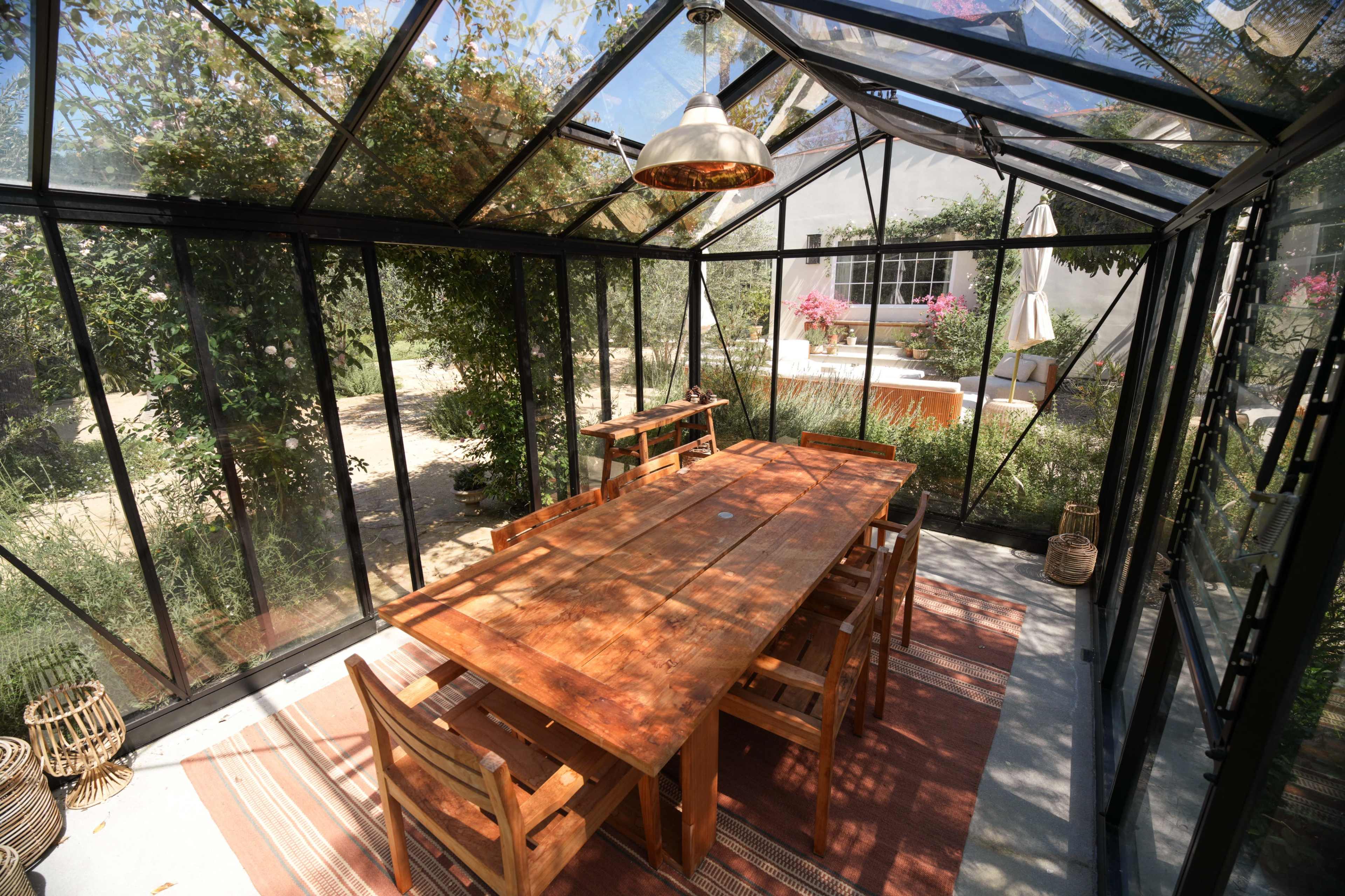 A spacious glass greenhouse features a large wooden dining table surrounded by chairs, with greenery visible outside.