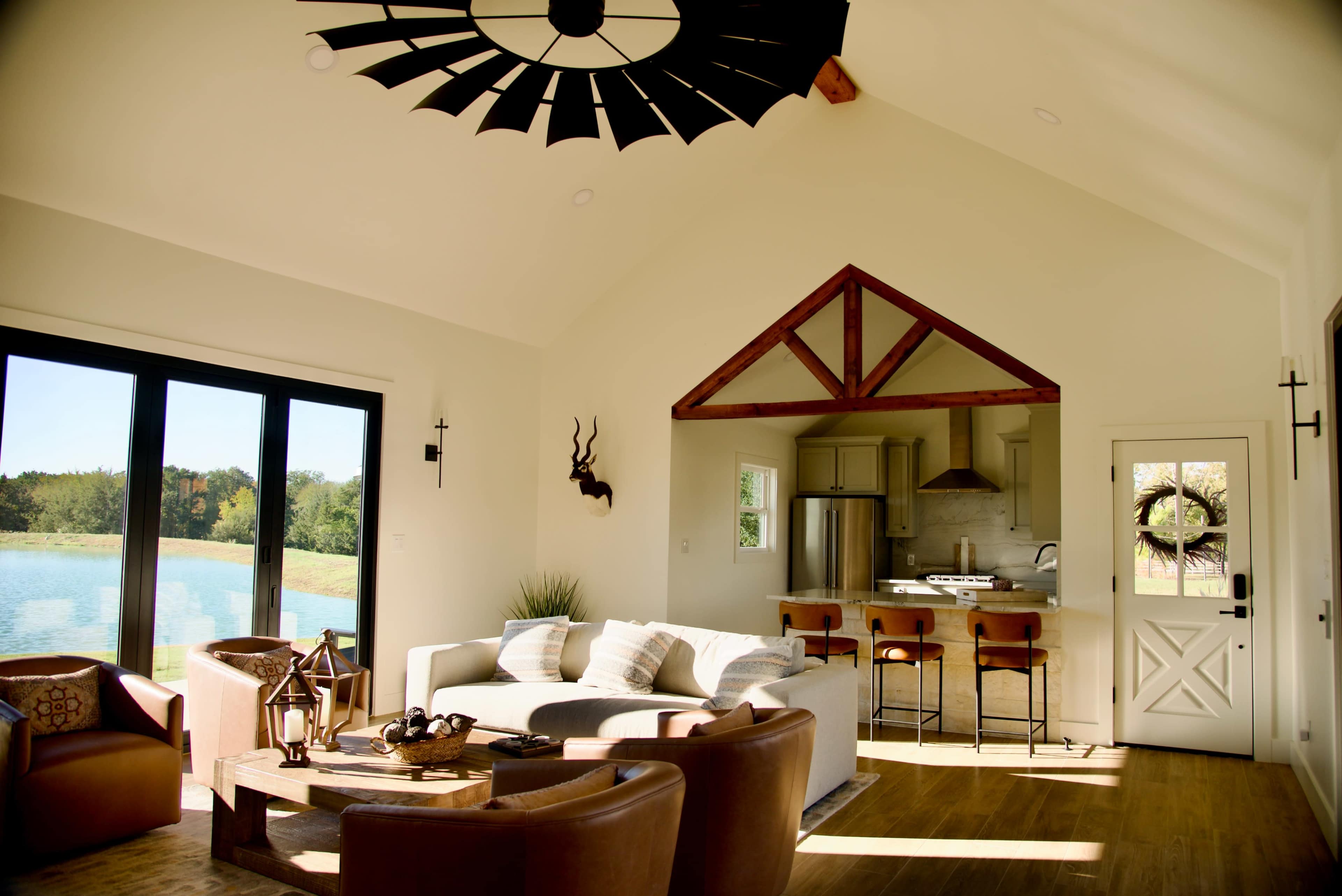 A spacious, modern living room features a tall ceiling, large windows overlooking a lake, and a kitchen area with wooden accents.