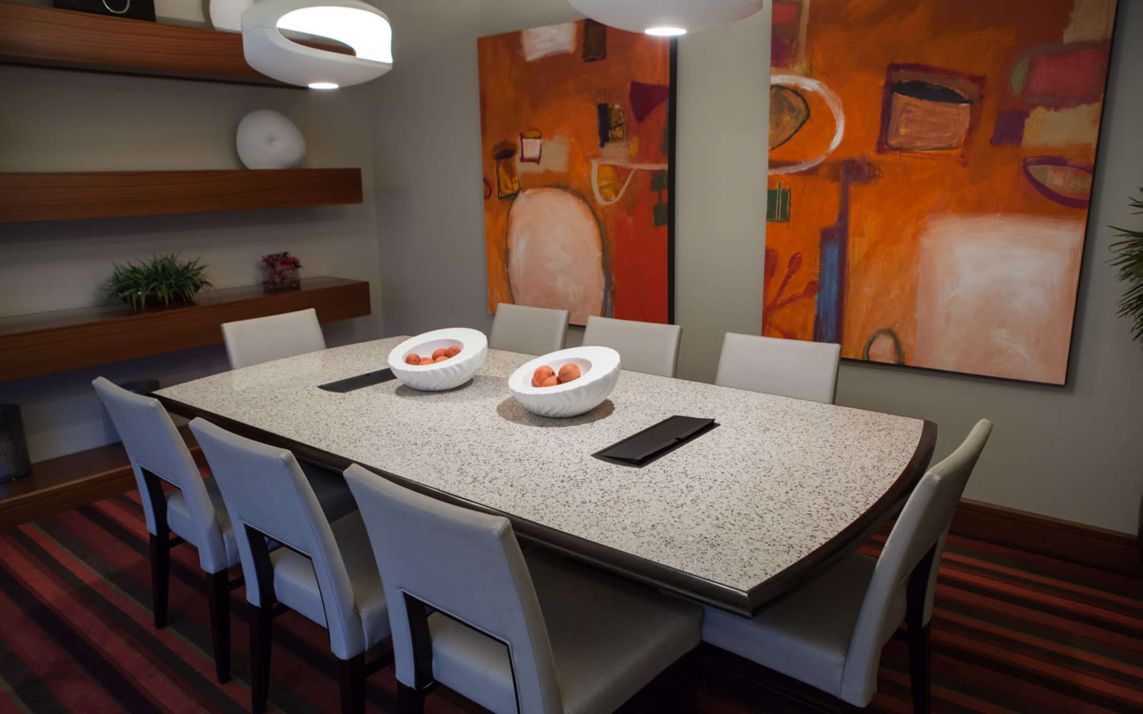 A modern dining room features a long table set with two bowls of fruit, surrounded by white chairs and flanked by colorful abstract artwork.