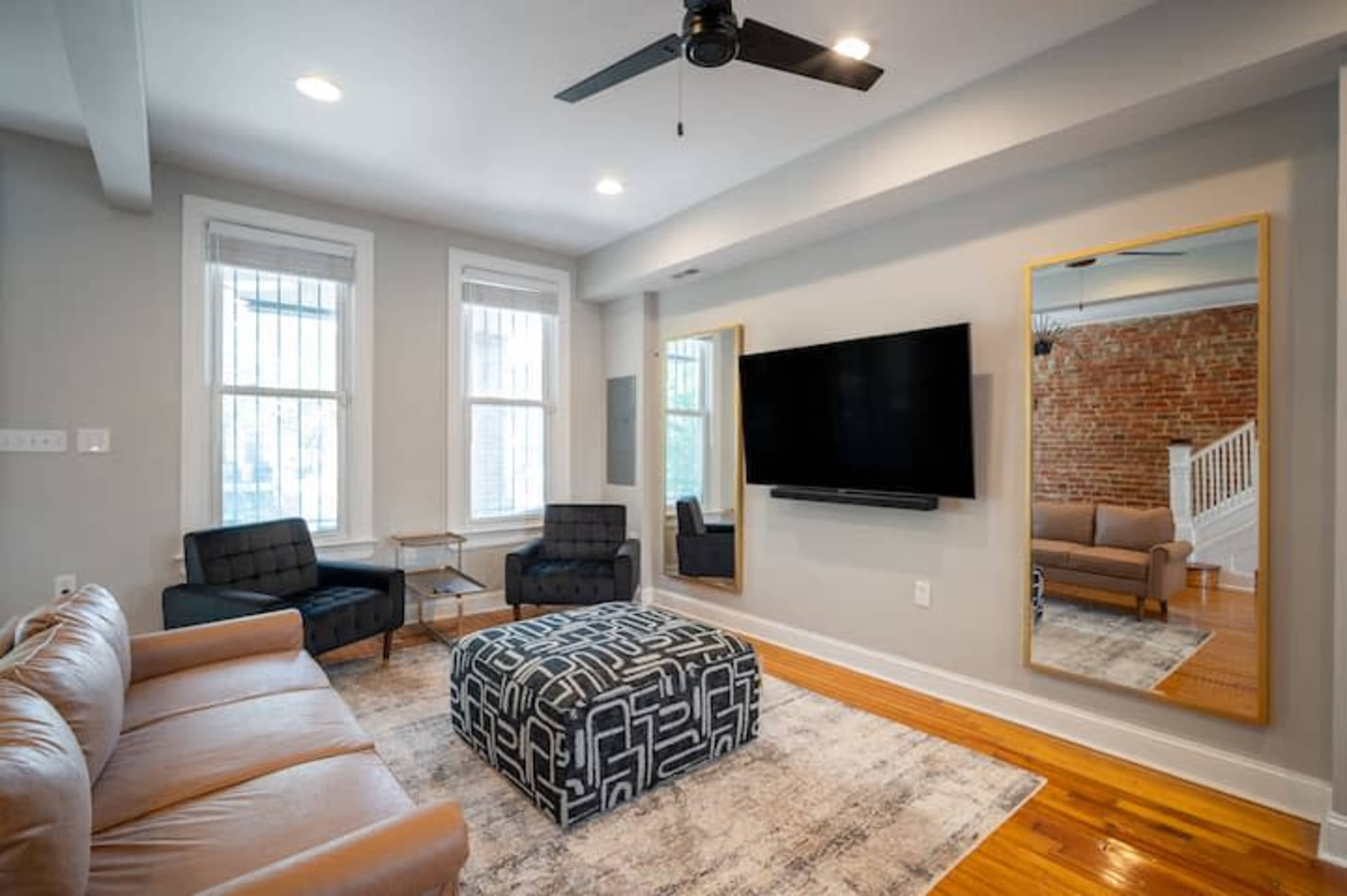 A living room with a large television mounted on a wall, a leather sofa, two black armchairs, a patterned ottoman, and large mirrors on either side.
