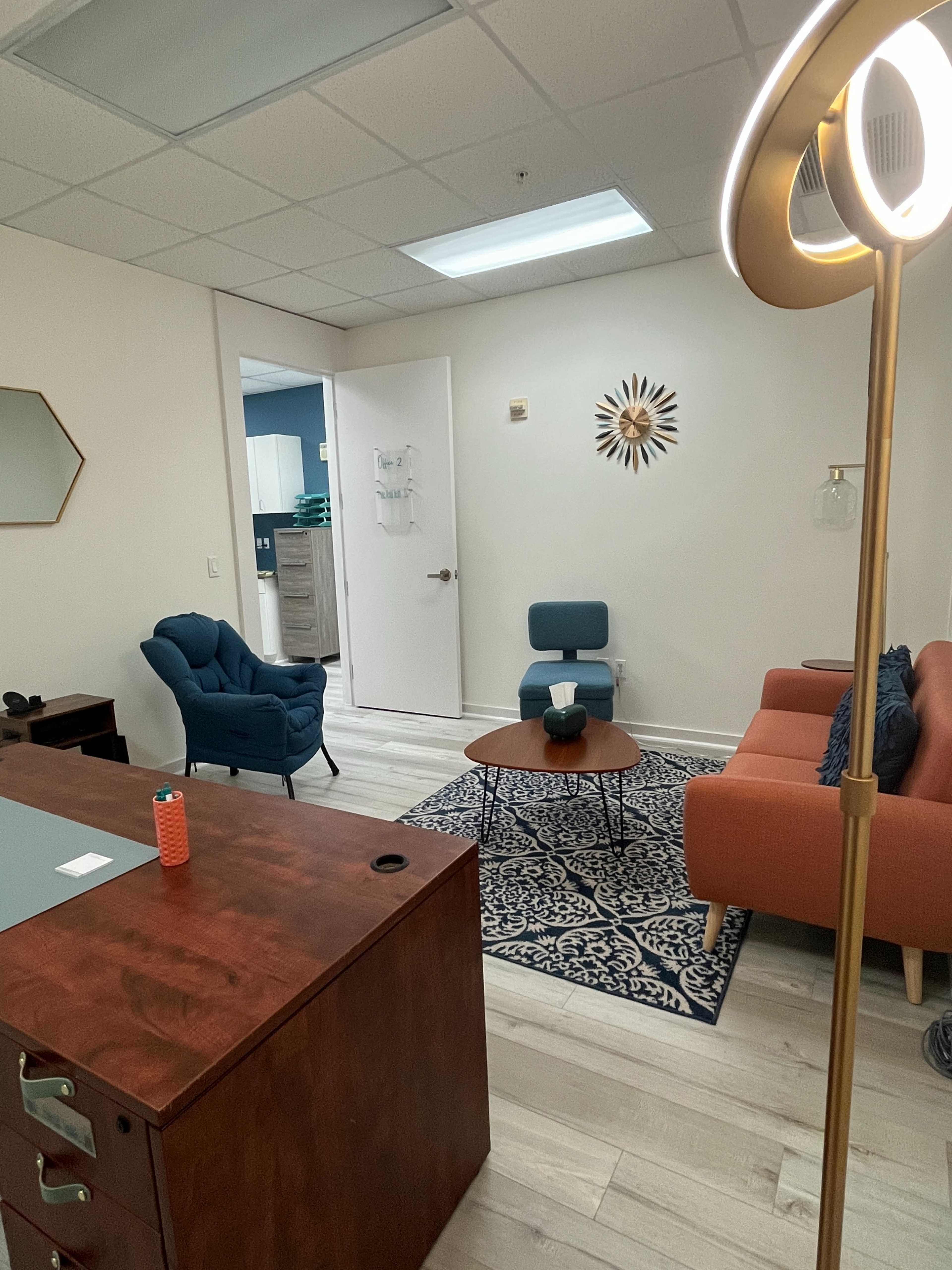 A small office space with a wooden desk, an orange sofa, a blue chair, a round coffee table, and a patterned rug, along with a visible kitchenette through a door.