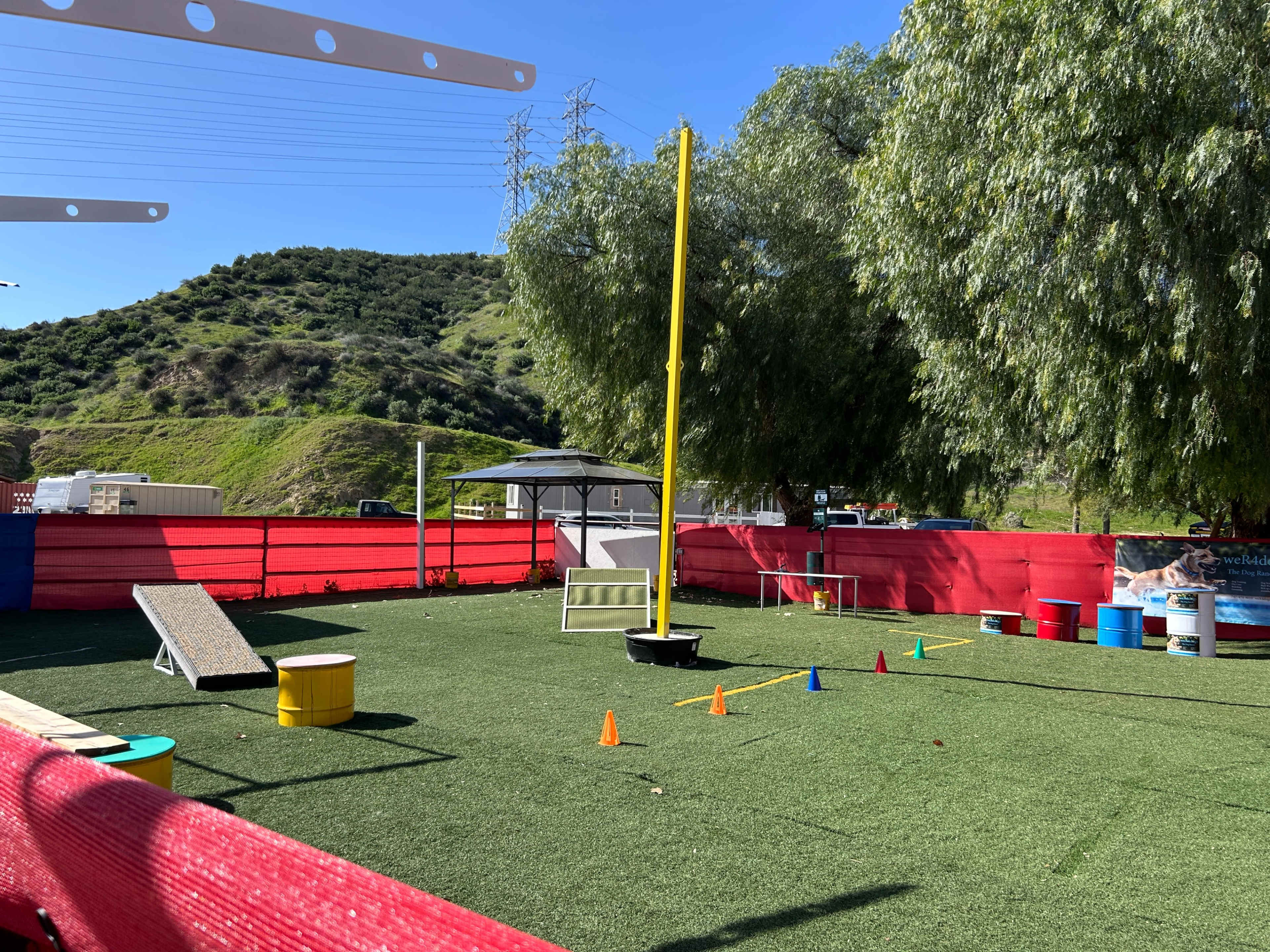 The image shows an outdoor training area with various obstacles, including cones, a ramp, and a tall yellow pole, surrounded by red barriers and green grass-like turf.
