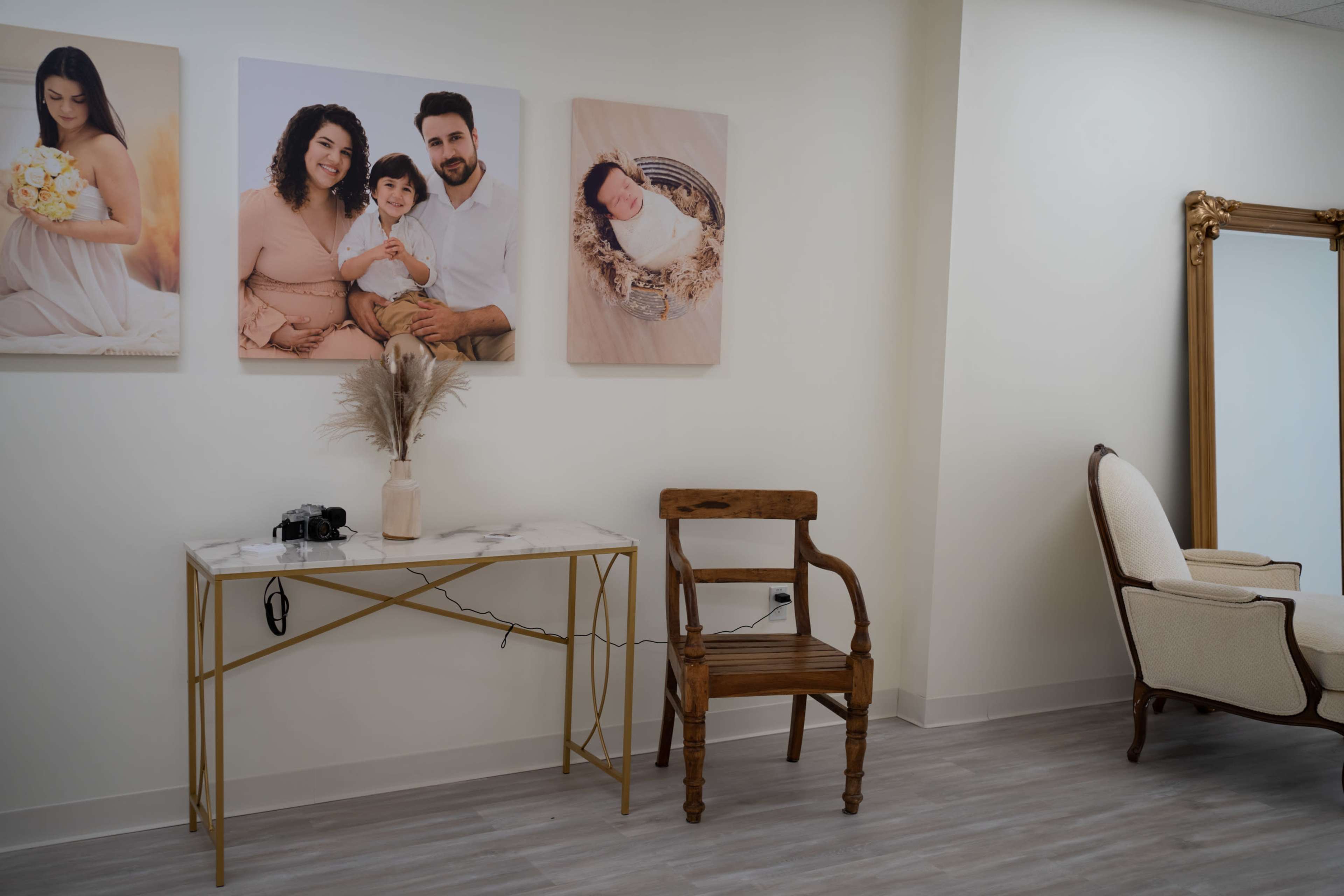 A room with light-colored walls featuring family portraits, a marble-topped table with a camera, a wooden chair, and an elegant armchair near a large mirror.