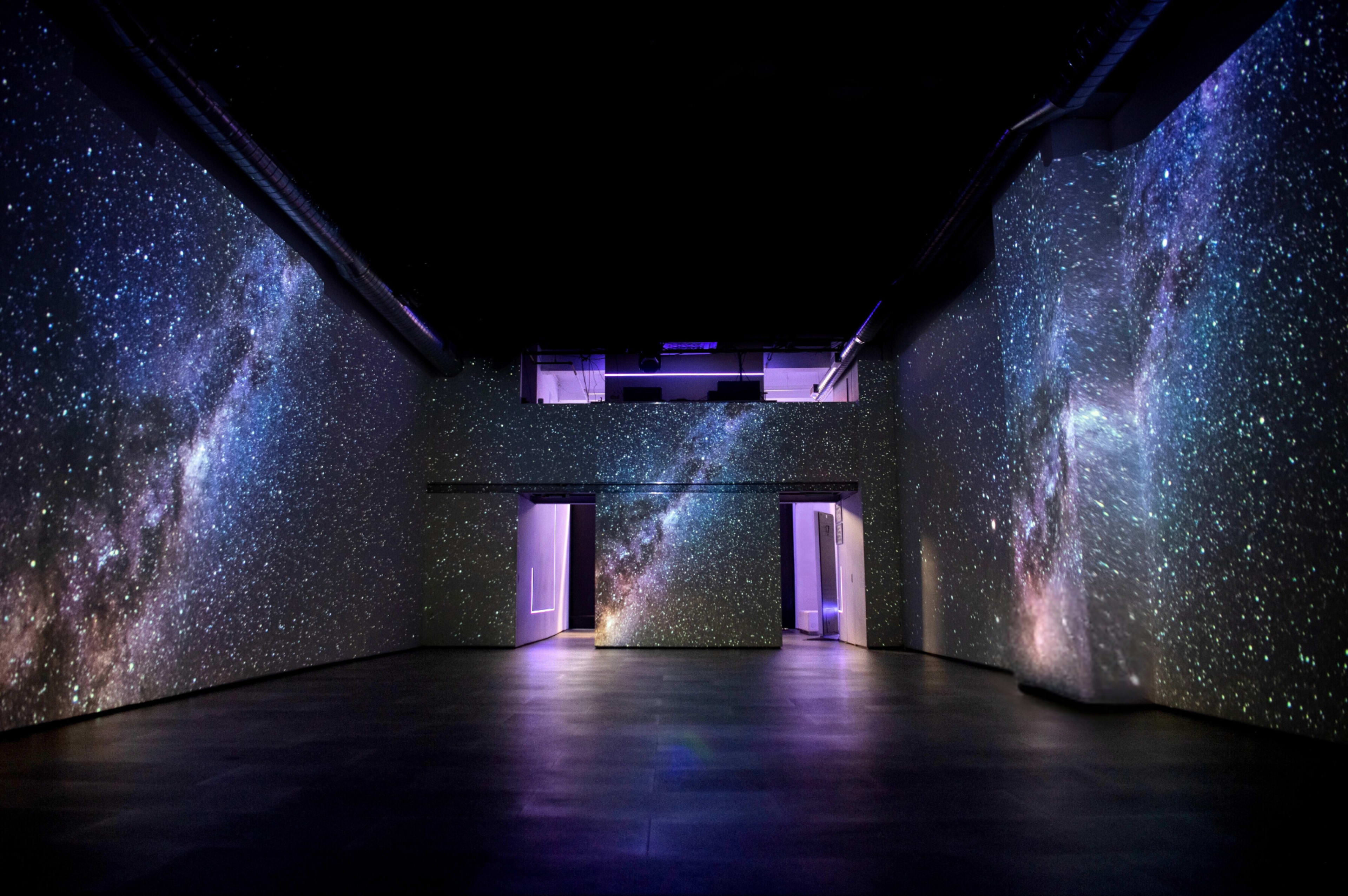 The image shows an art installation in a large, dark room with projections of a starry galaxy covering the walls.