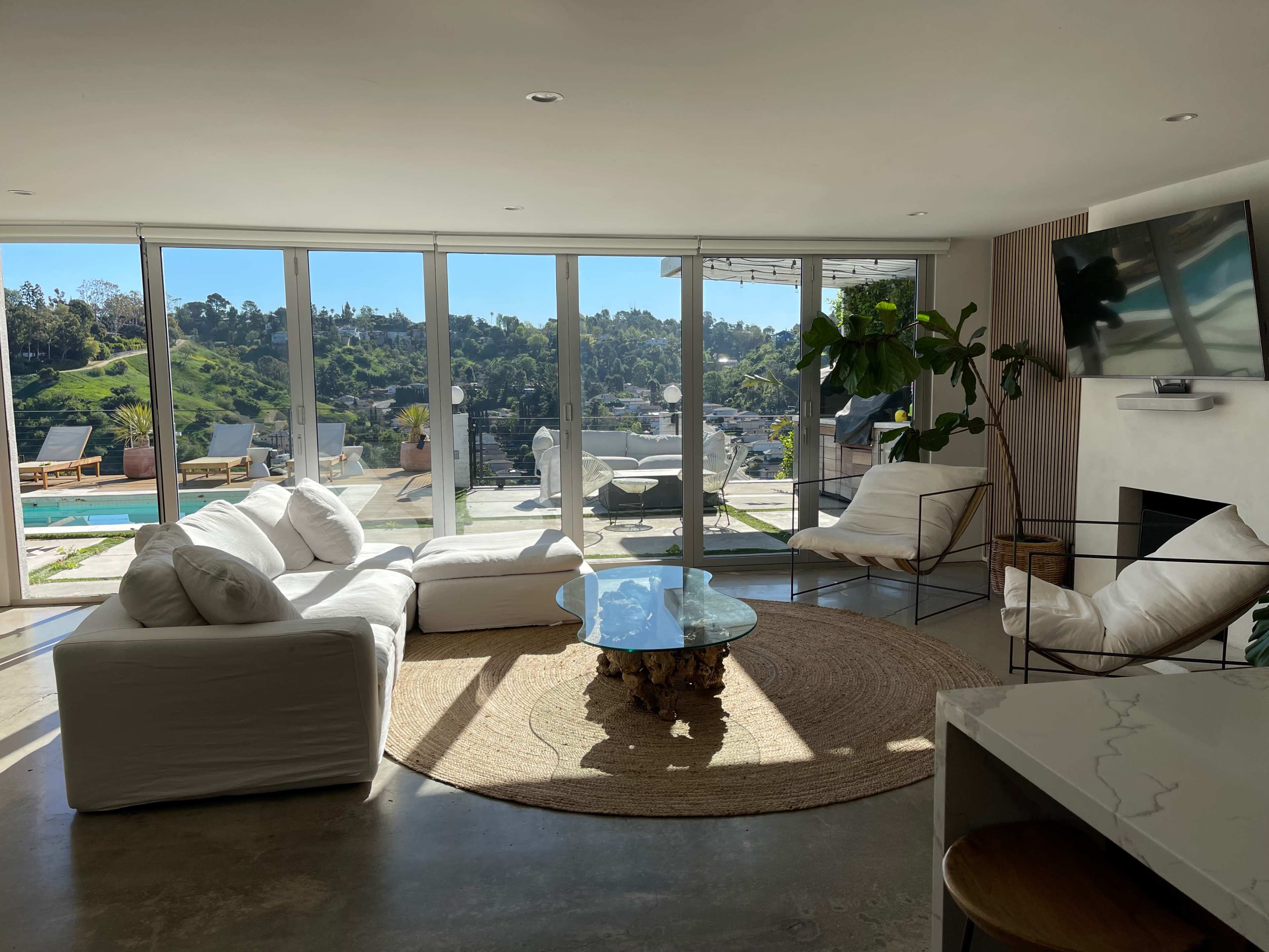 The living room features a white sectional sofa, two chairs, a glass coffee table, and large windows displaying a view of a hillside and pool area.