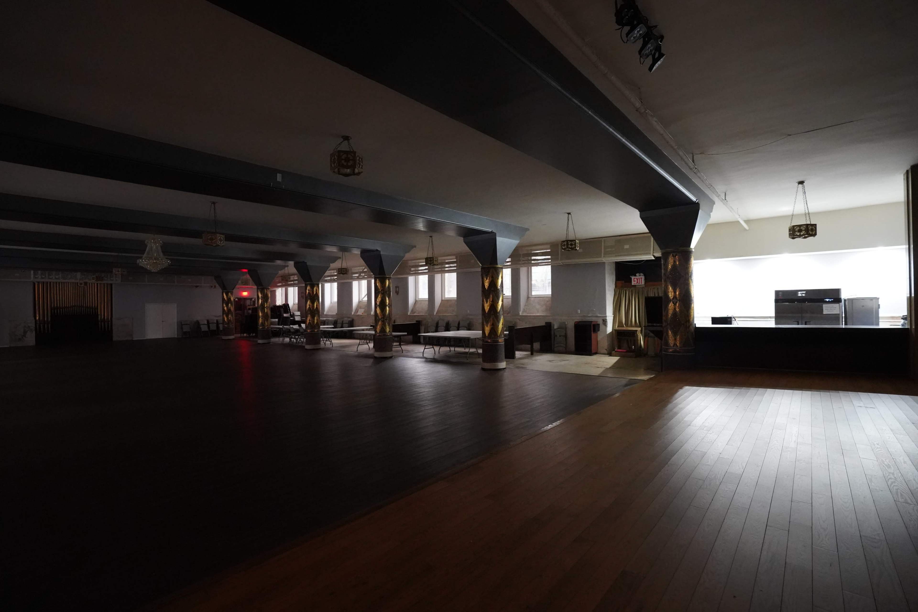 An empty event space with wooden flooring, tall windows, and dark pillars.