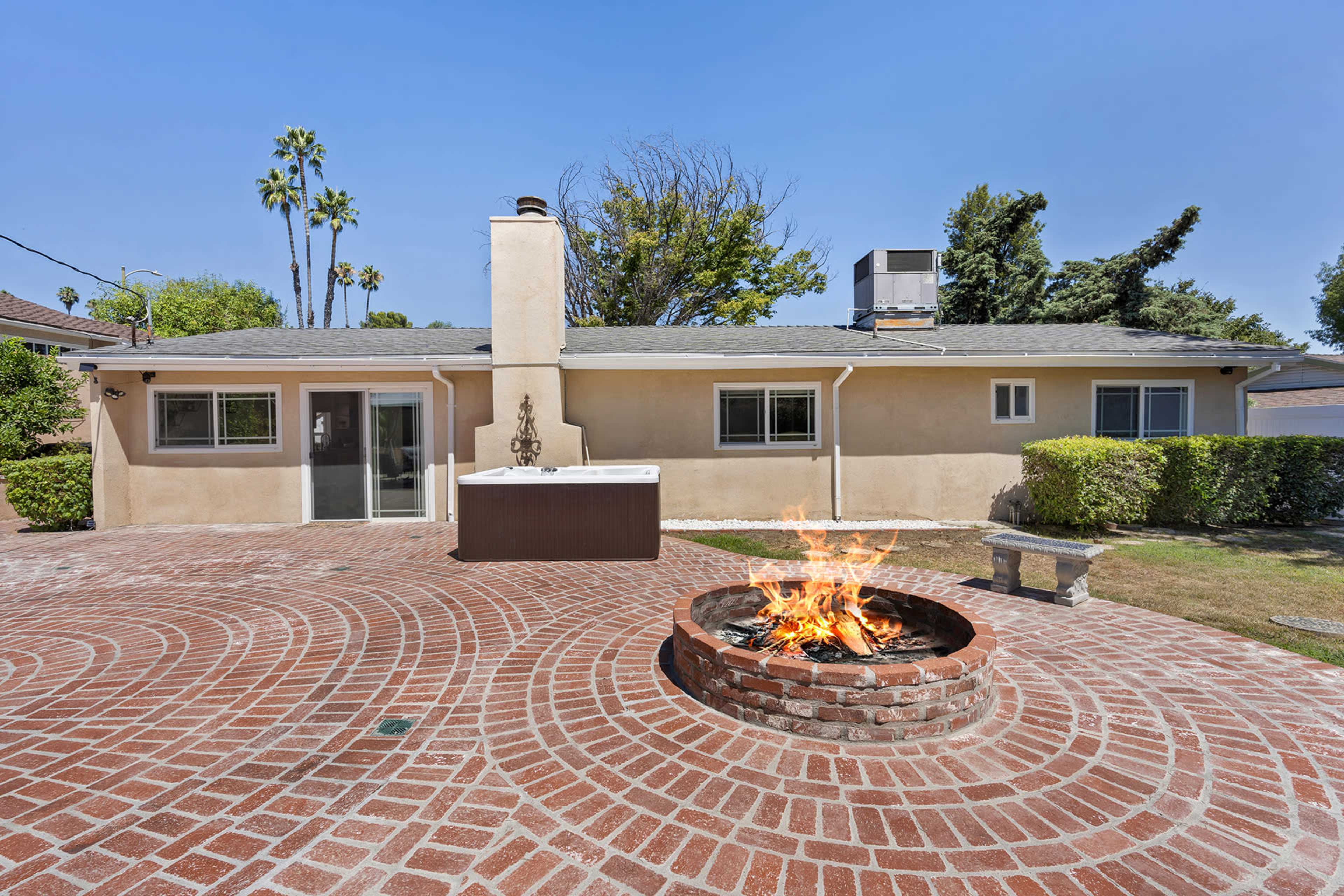 A brick patio with a fire pit is situated in front of a single-story house surrounded by greenery.