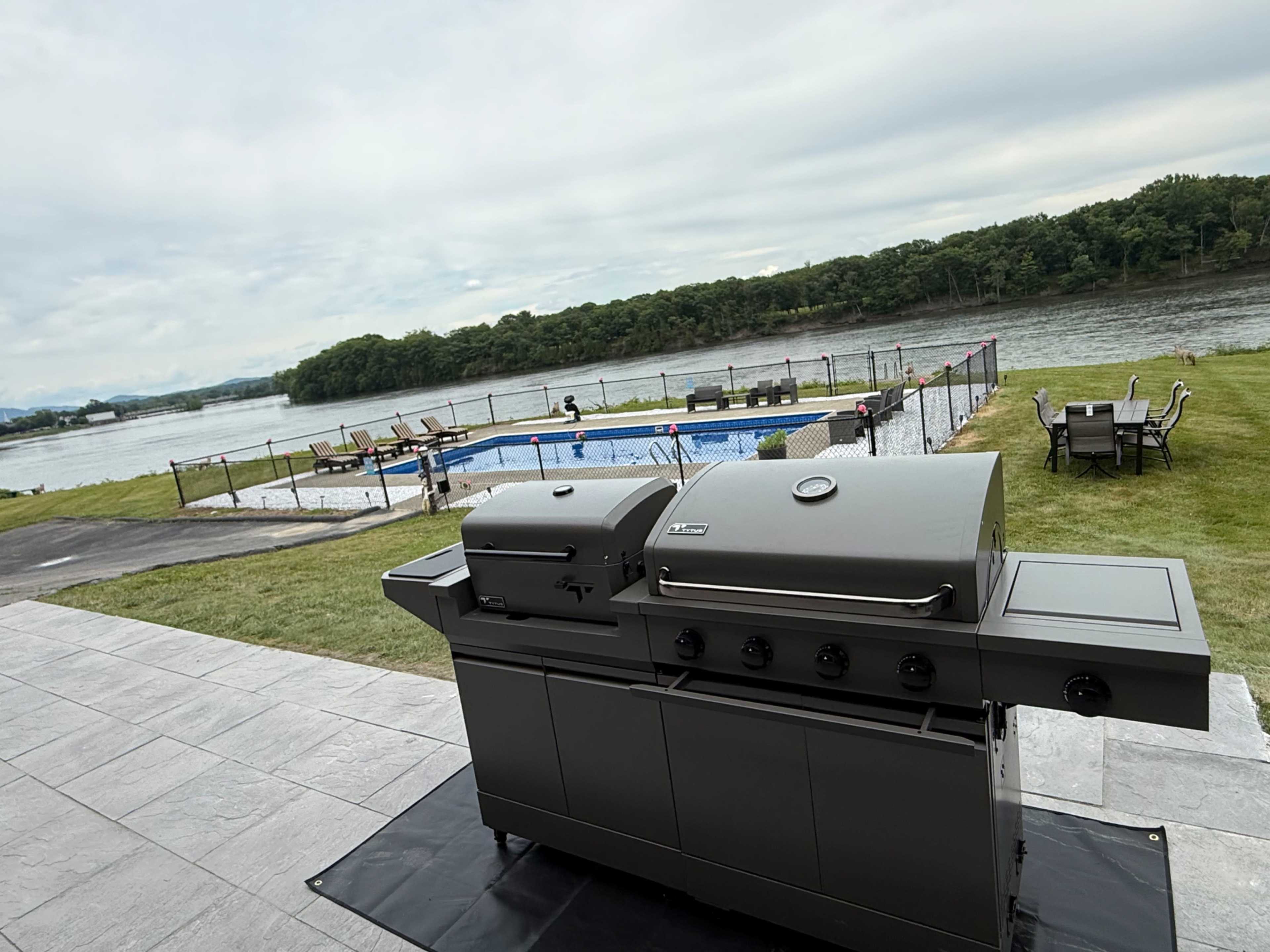 A modern grill is positioned on a patio overlooking a pool and a river, with chairs and lounge areas nearby.