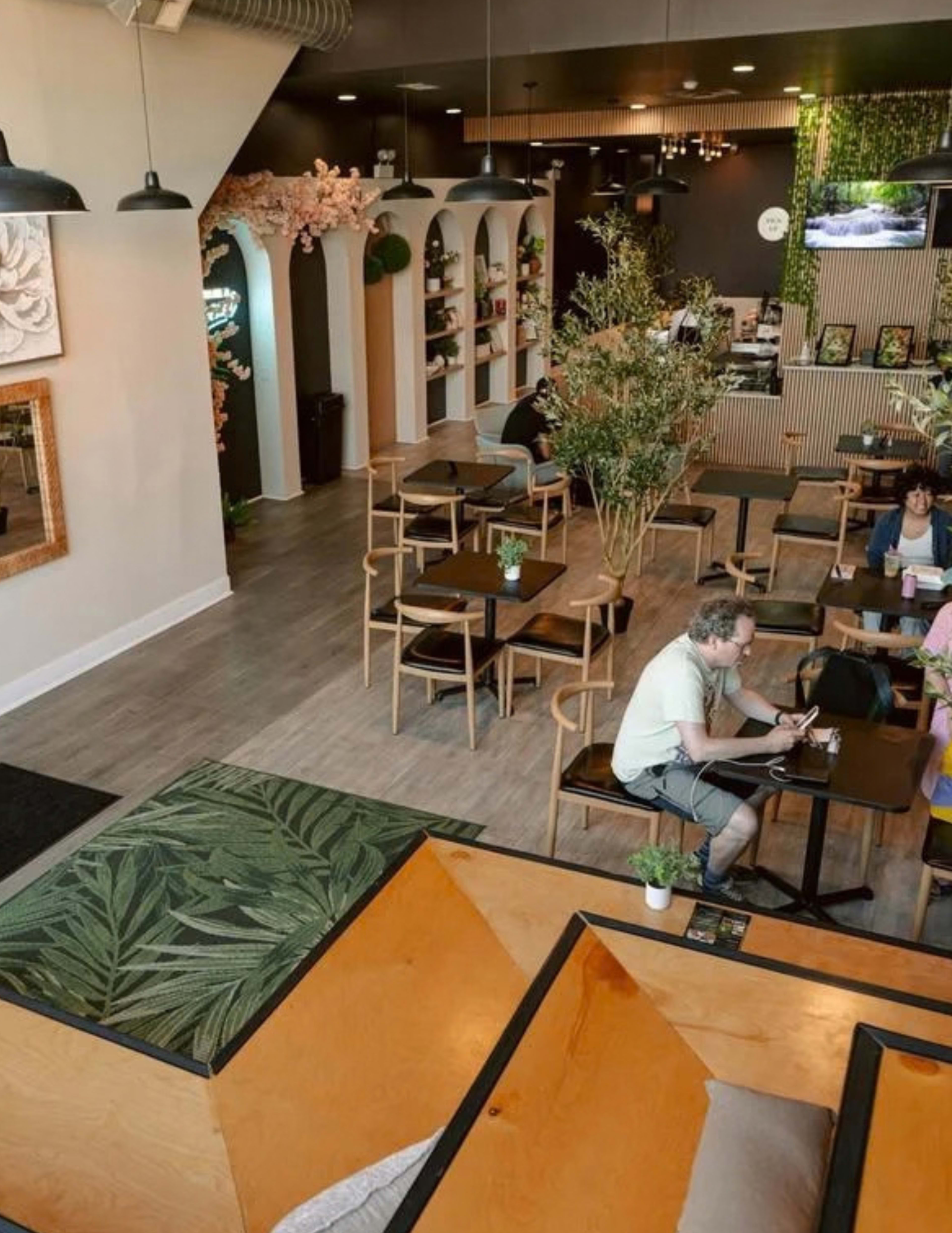 The image shows a modern café interior with wooden tables, green decor, and customers sitting and working at various seating areas.