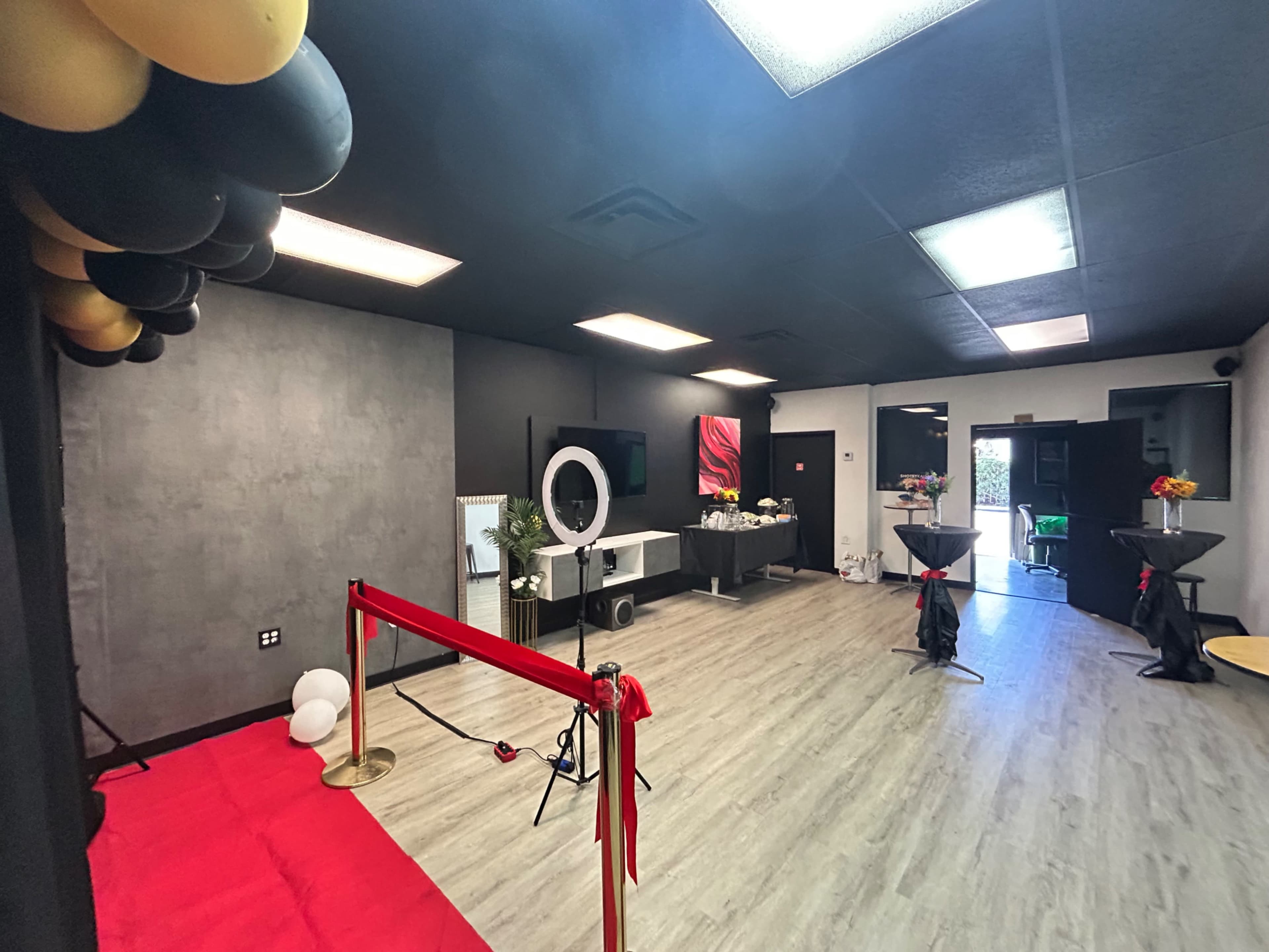 A spacious event room features a red carpet, black and gold balloons, a ring light setup, and several high-top tables arranged for an occasion.