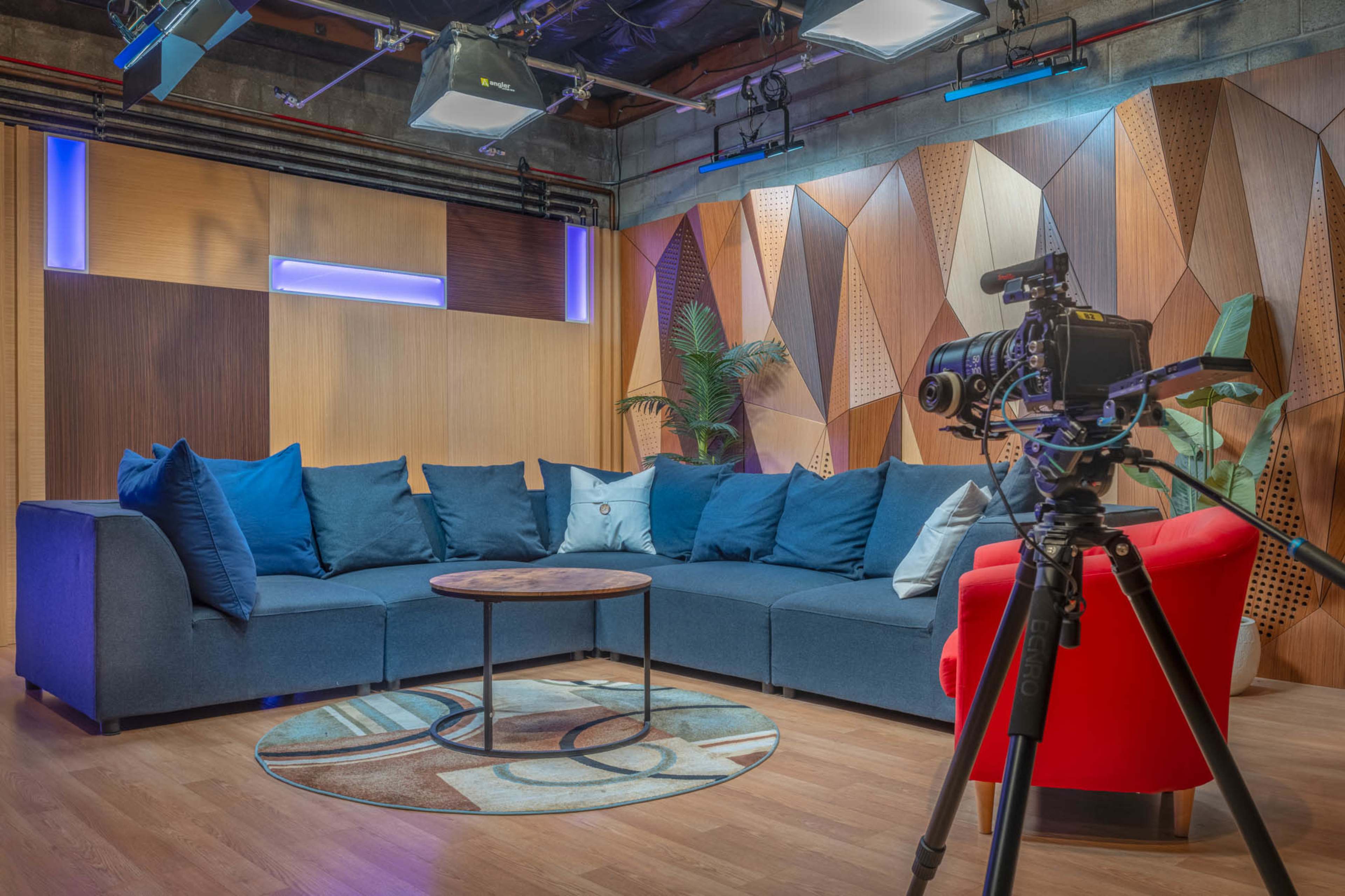 A modern studio set with a blue sofa, a round coffee table, and a camera on a tripod, surrounded by geometric wooden panels and accent lighting.