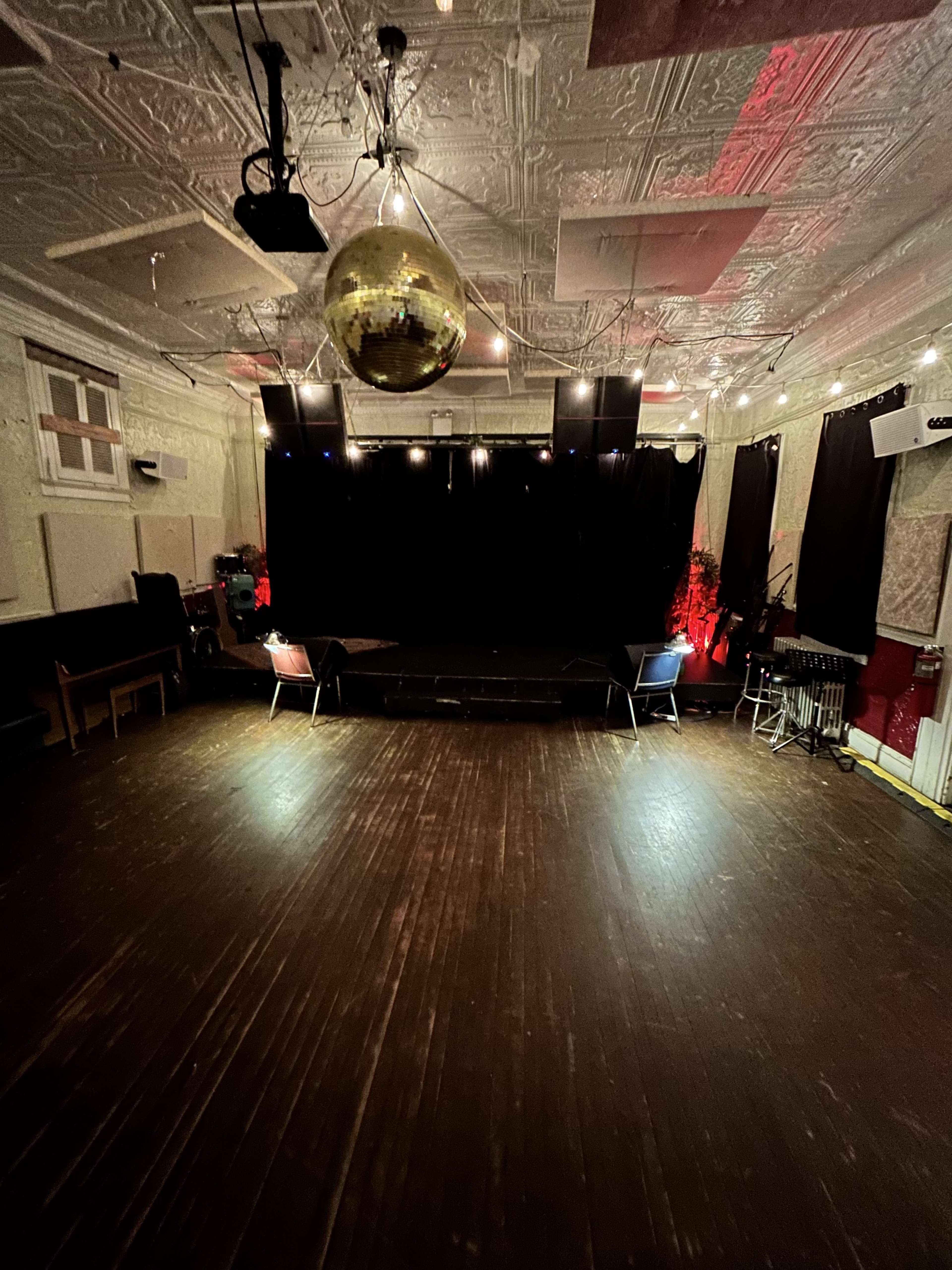 A dimly lit performance space features a raised stage with a black curtain, a disco ball overhead, and wooden floors.