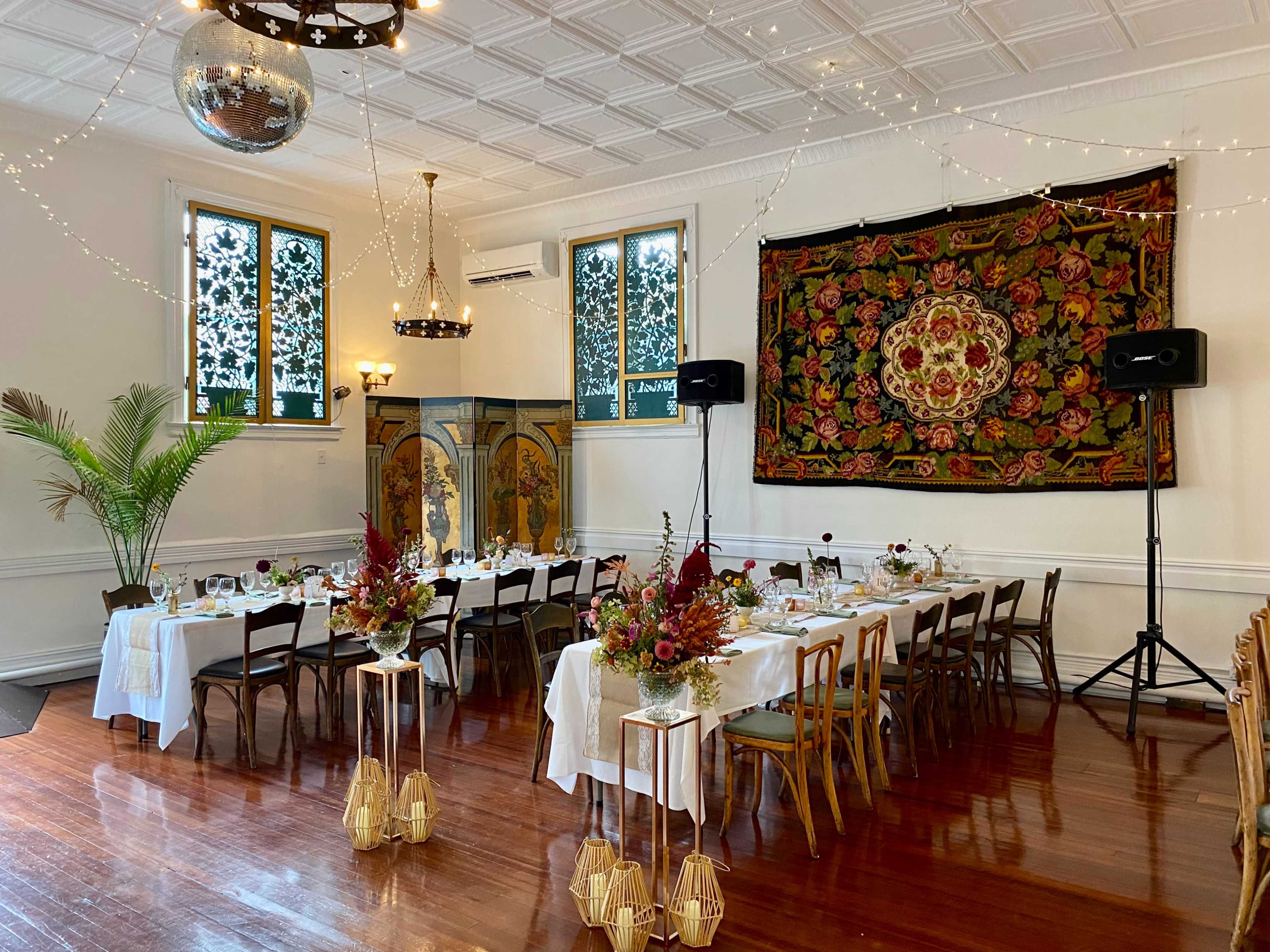 A dining area is set with multiple tables adorned with white tablecloths and floral centerpieces, featuring large windows and decorative elements on the walls.