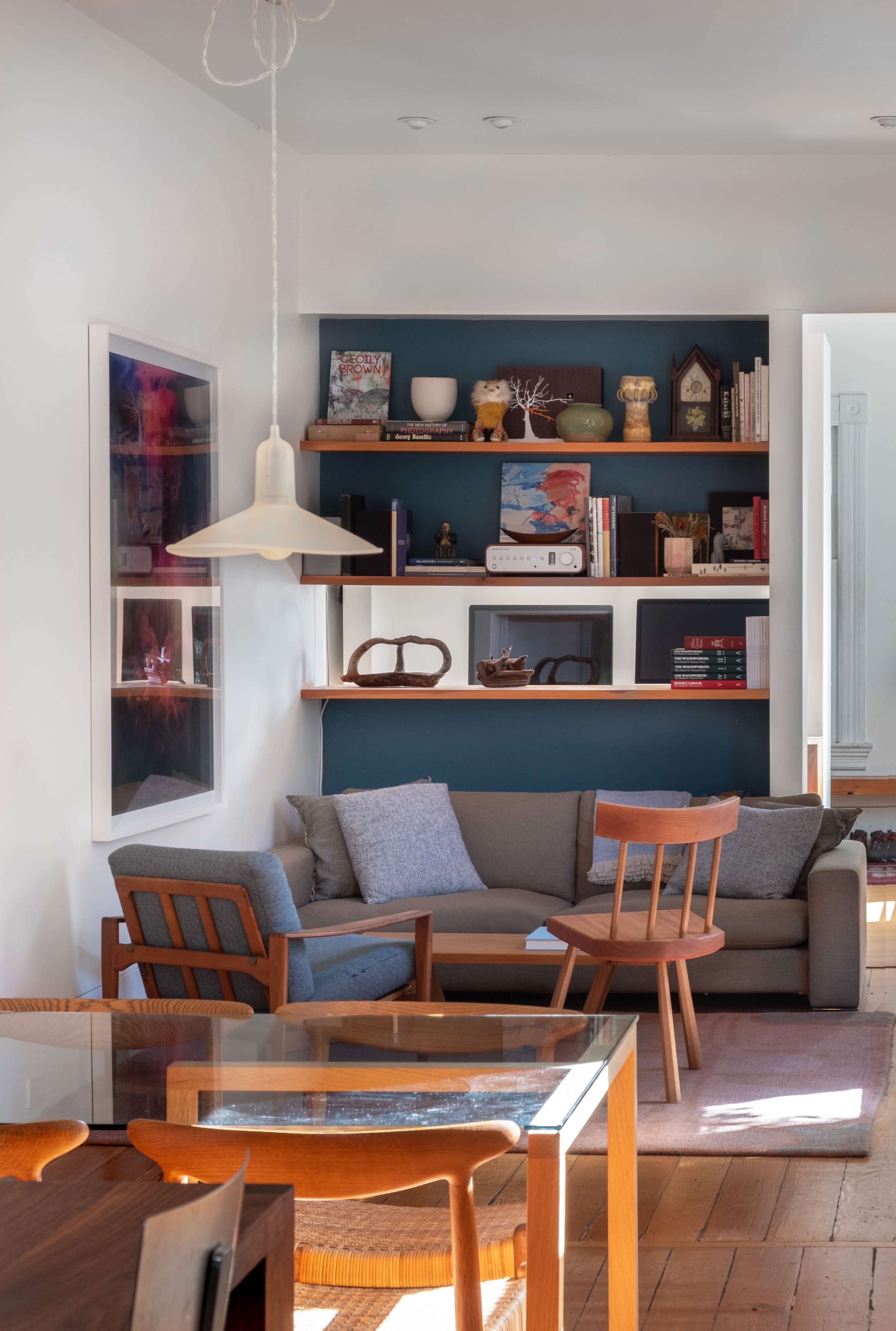 A modern living room features a gray couch with a pillow, wooden chairs, and a wall shelf displaying books and decor items.