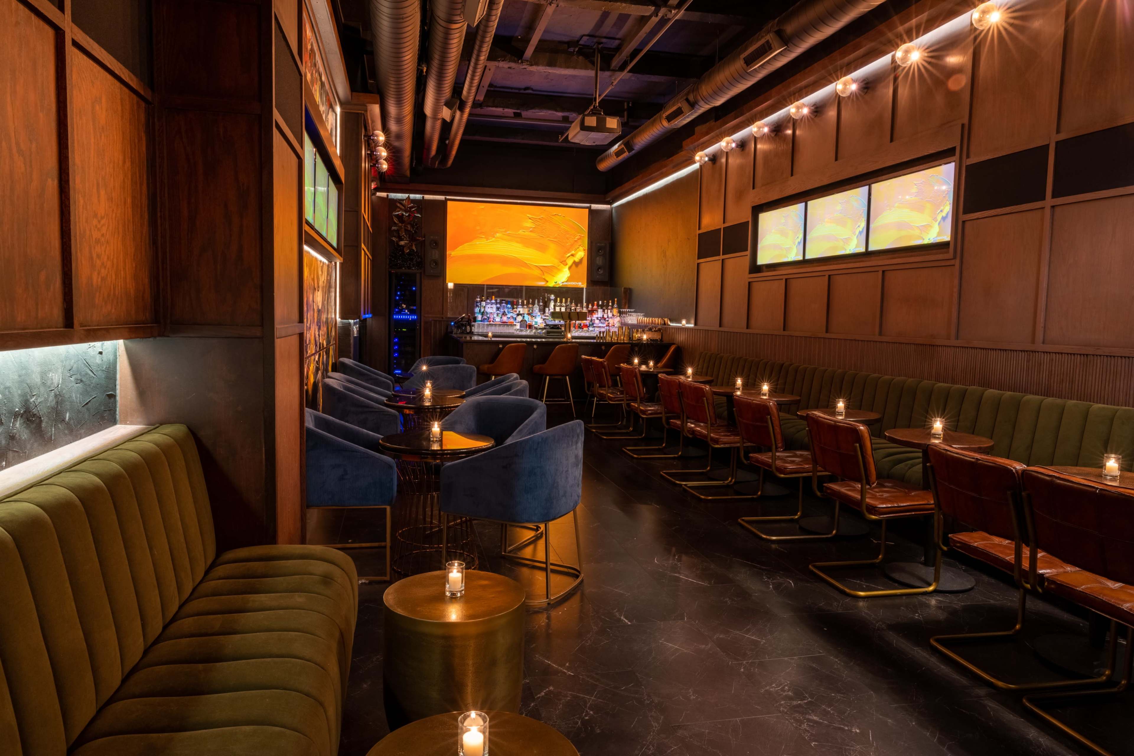 The image shows a stylish bar interior with a mix of green velvet and blue upholstered seating, dark wood paneling, and dim lighting cast by candles on small tables.