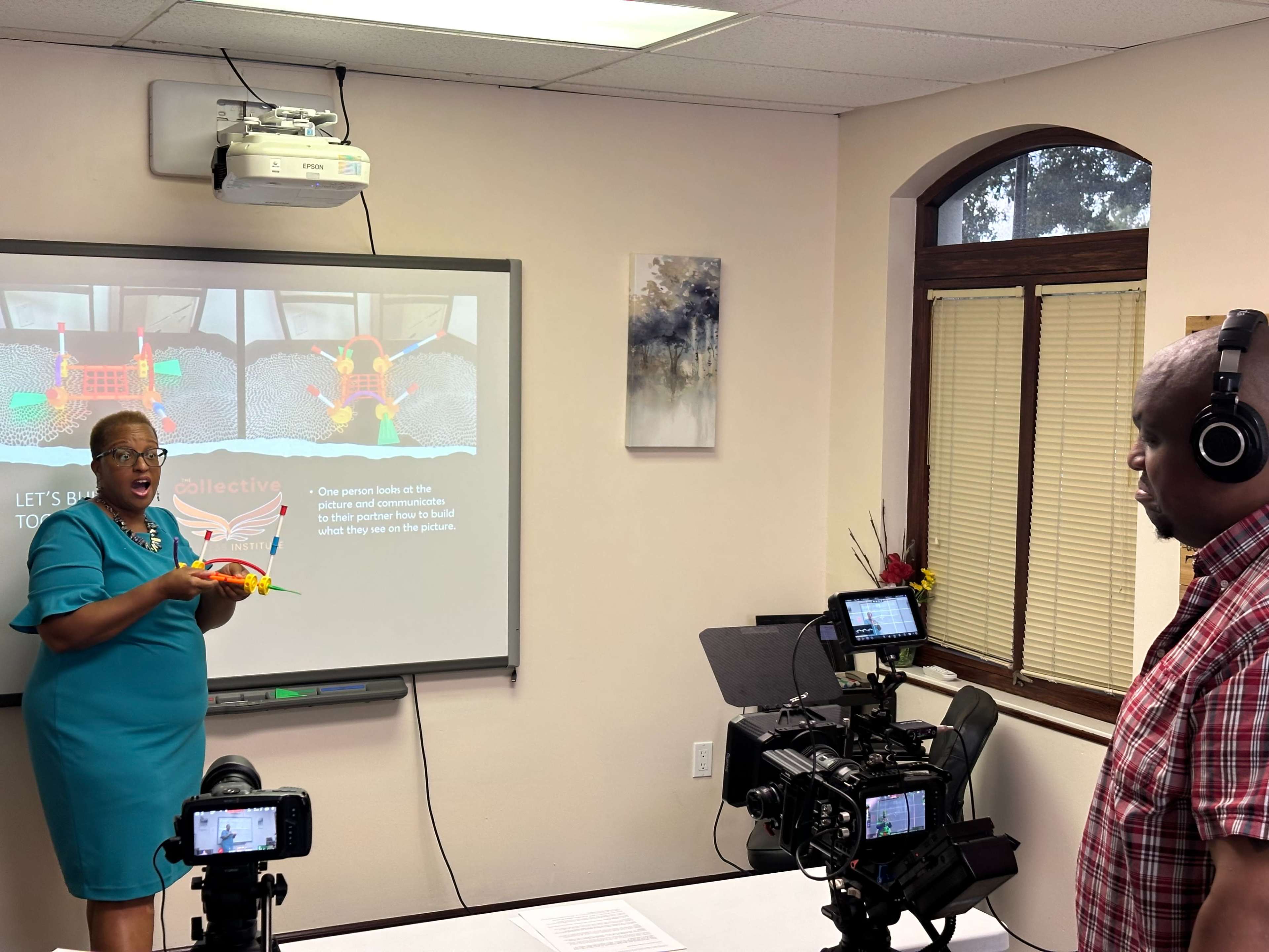 A woman demonstrates a project using a toy in front of a presentation screen while a man listens and records the session with a video camera.