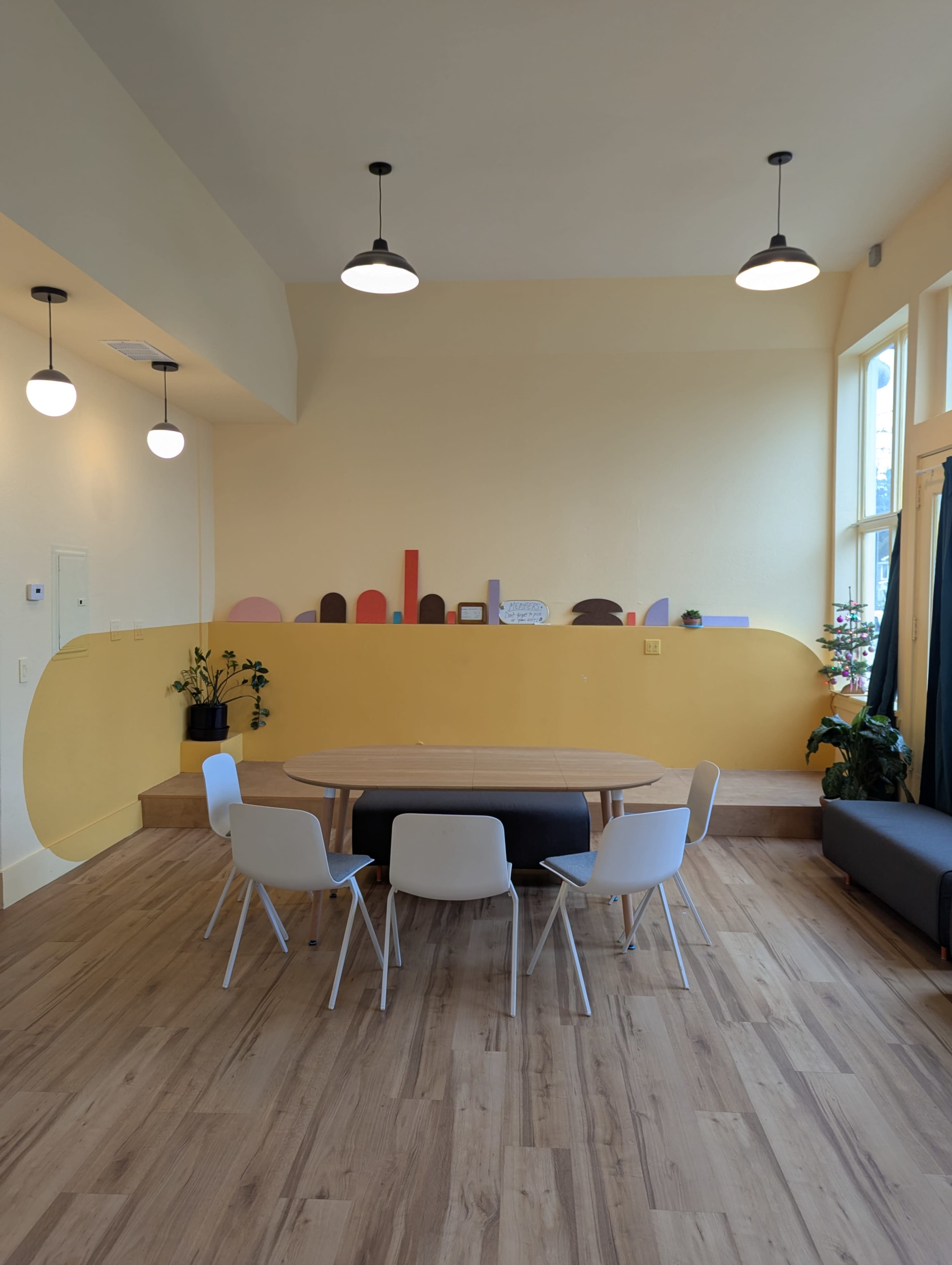 The image shows a bright room with a round wooden table surrounded by white chairs, a yellow wall with abstract cutouts, and large windows letting in natural light.