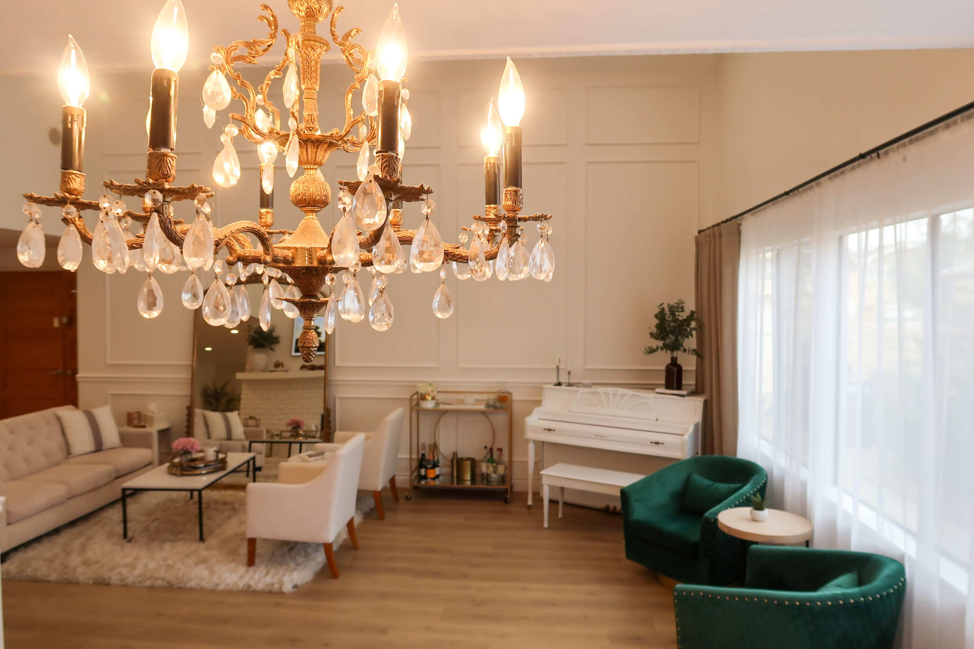 A chandelier hangs prominently from the ceiling above a bright living area featuring a sofa set, two green chairs, and a white piano against the wall.